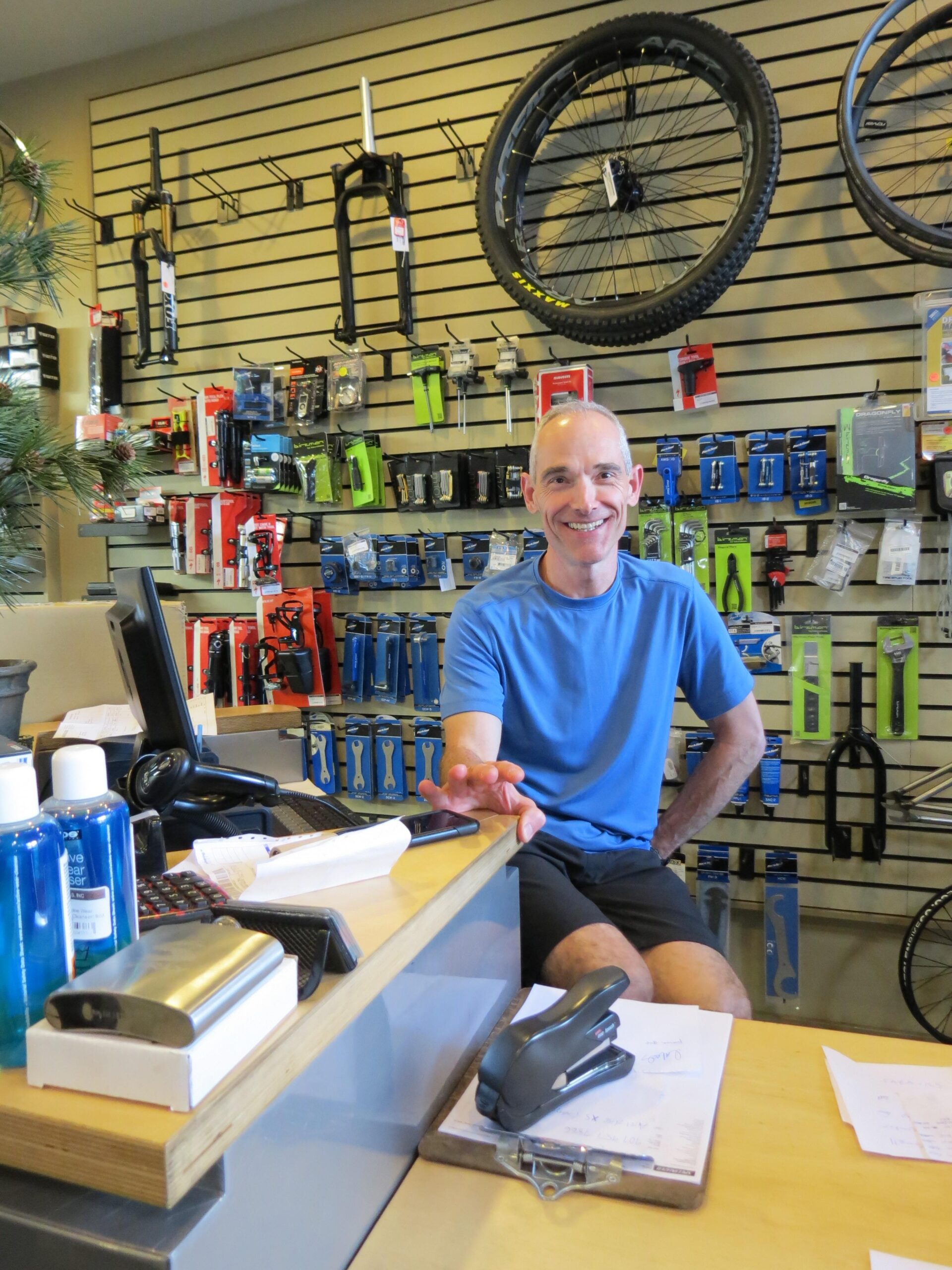 A friendly employee sitting at a counter in a bike shop, surrounded by various bicycle parts and accessories displayed on the wall behind him. He is wearing a blue t-shirt and shorts, smiling as he engages with customers. The counter features a computer, a stapler, and product displays.