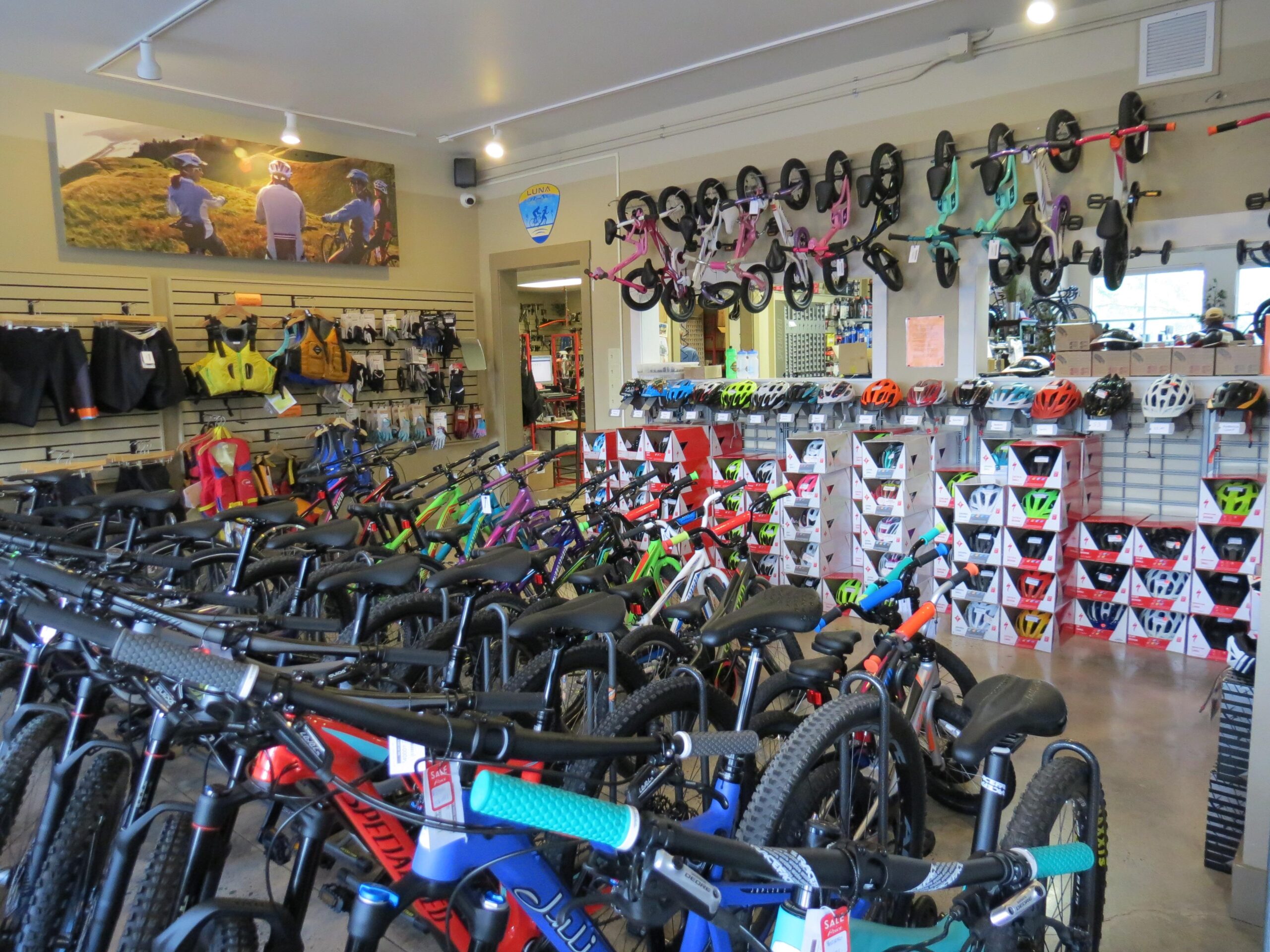 Interior view of a bike shop featuring a variety of bicycles, including mountain bikes, alongside an assortment of helmets and biking gear displayed on shelves. The shop is well-lit and organized, with bicycles lined up closely together and some children's balance bikes hung on the wall. In the background, there is a large photo mural depicting people cycling outdoors.