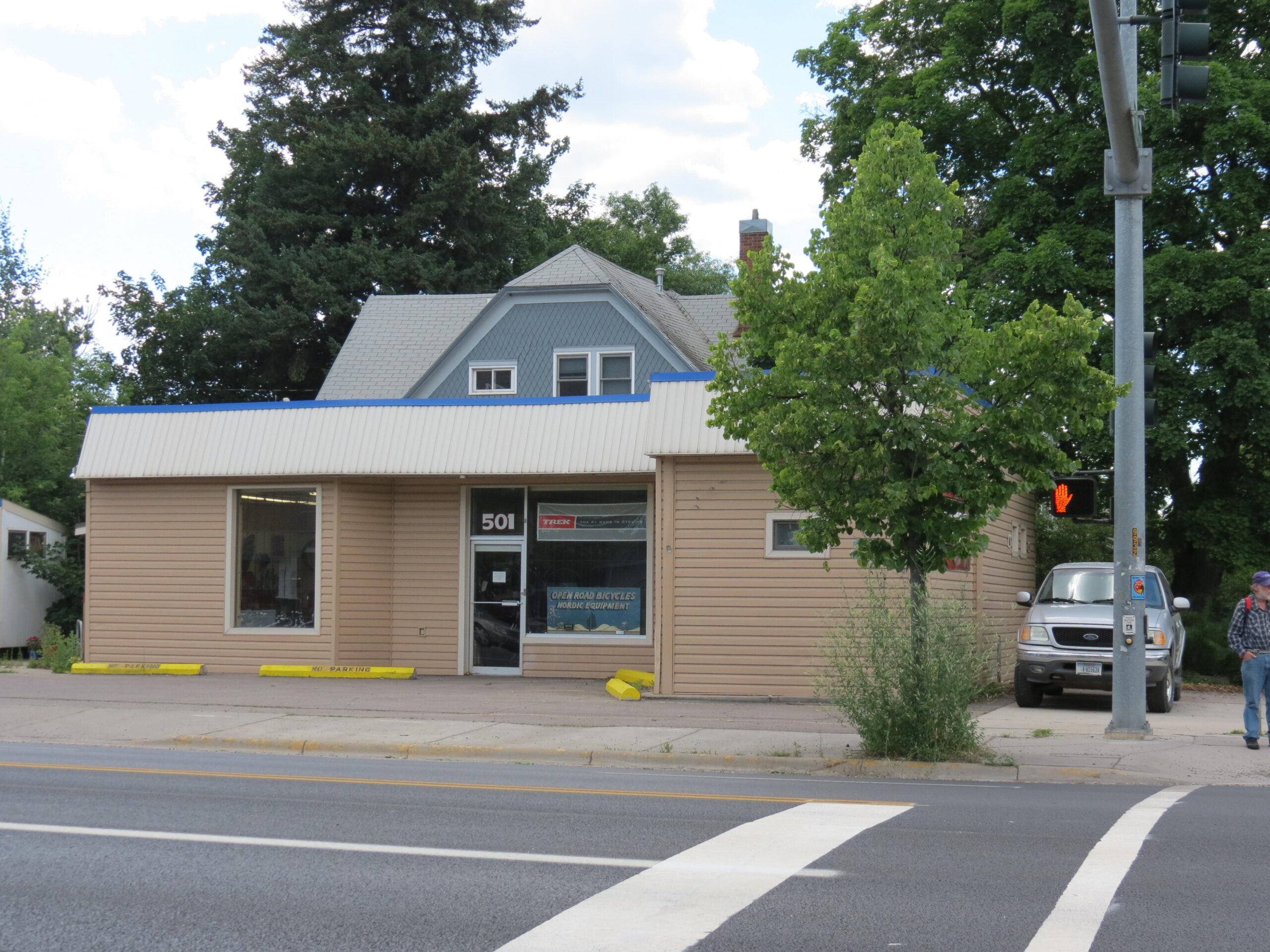 A small commercial building with beige siding and a blue, sloped roof. The front features large windows, including a door marked with the number "501." Yellow markings indicate no parking in front of the building. A tree is positioned outside, and a parked car is visible on the side. In the foreground, a person is seen walking along the sidewalk. Traffic light signals are visible nearby.