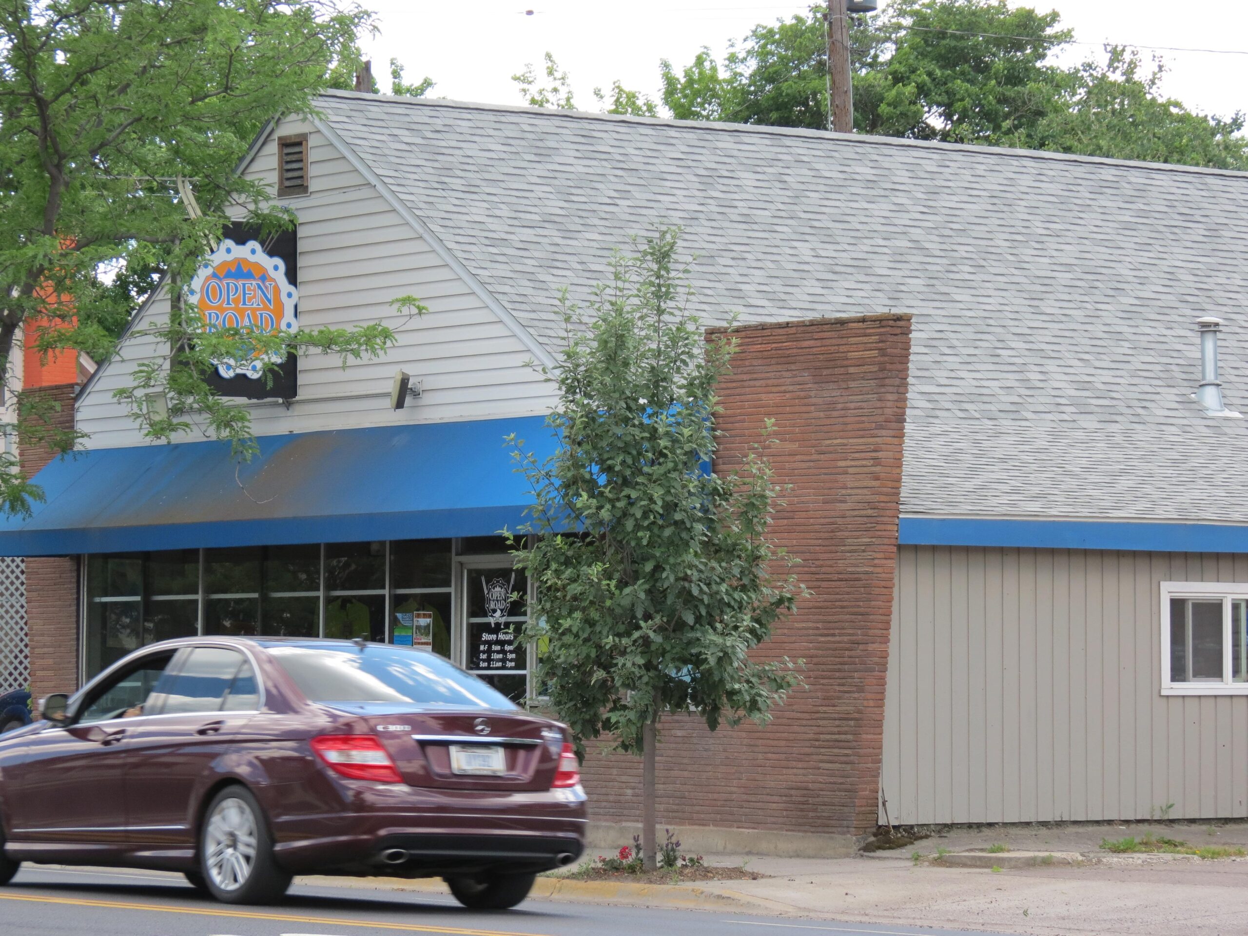 Image of a small building with a sign reading "OPEN ROAD." The structure has a gray roof, blue awning, and large windows. A red car is parked in front, driving on a road lined with greenery and trees in the background.
