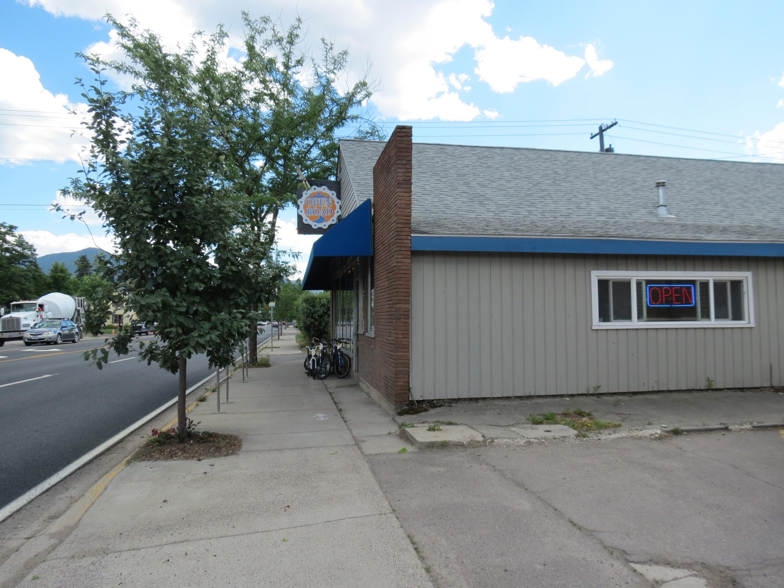 A corner view of a small building with a blue awning and a red "OPEN" neon sign in the window. A tree is visible in front of the building, and several bicycles are parked nearby. The scene is set along a busy street with vehicles passing by, under a partly cloudy sky.