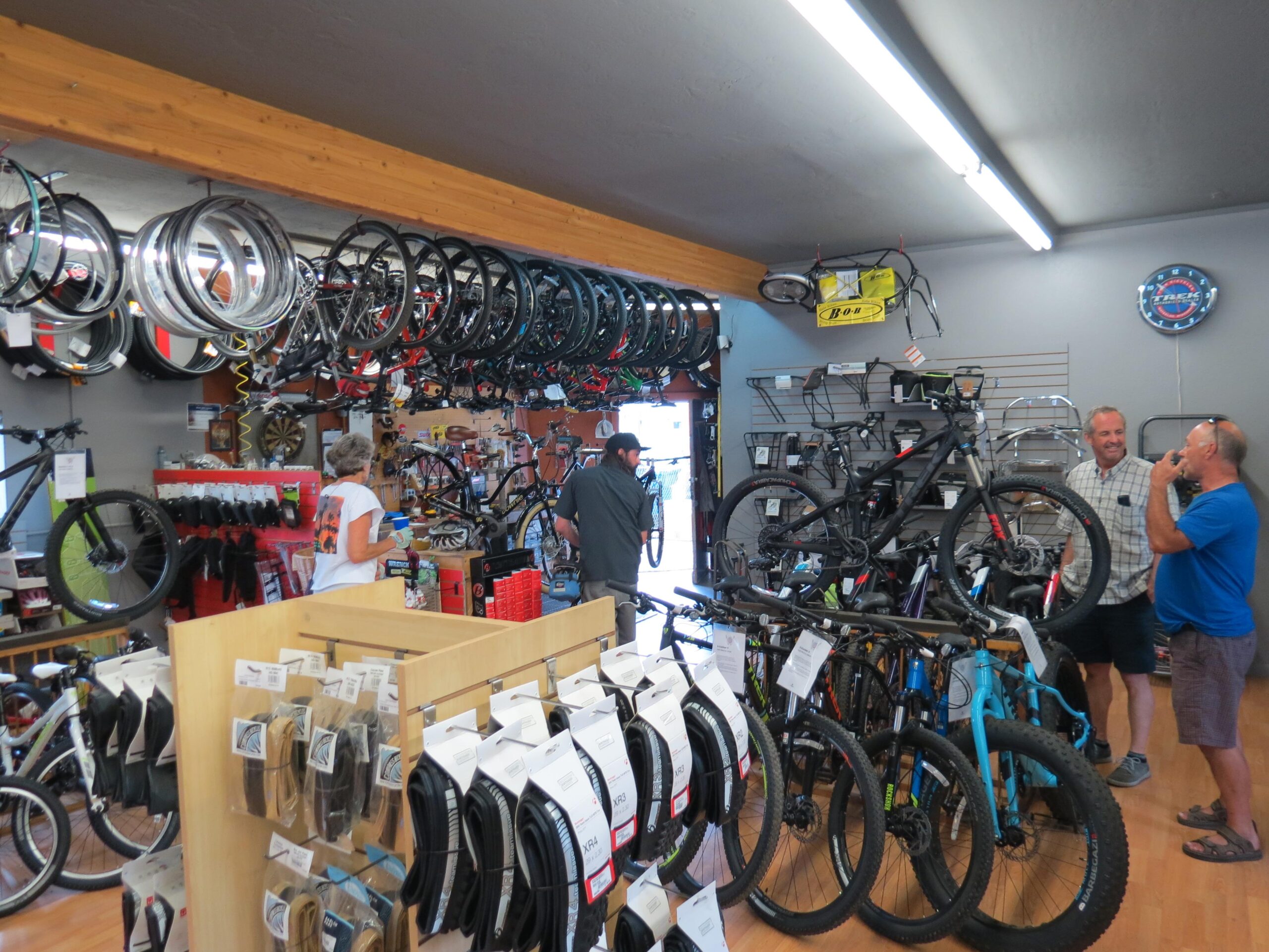 A bustling bicycle shop interior, featuring a display of various bike models, accessories, and tires. Customers engage in conversation while exploring the merchandise. The shop is well-lit, with bicycles hanging from the ceiling, showcasing a vibrant selection of cycling products.