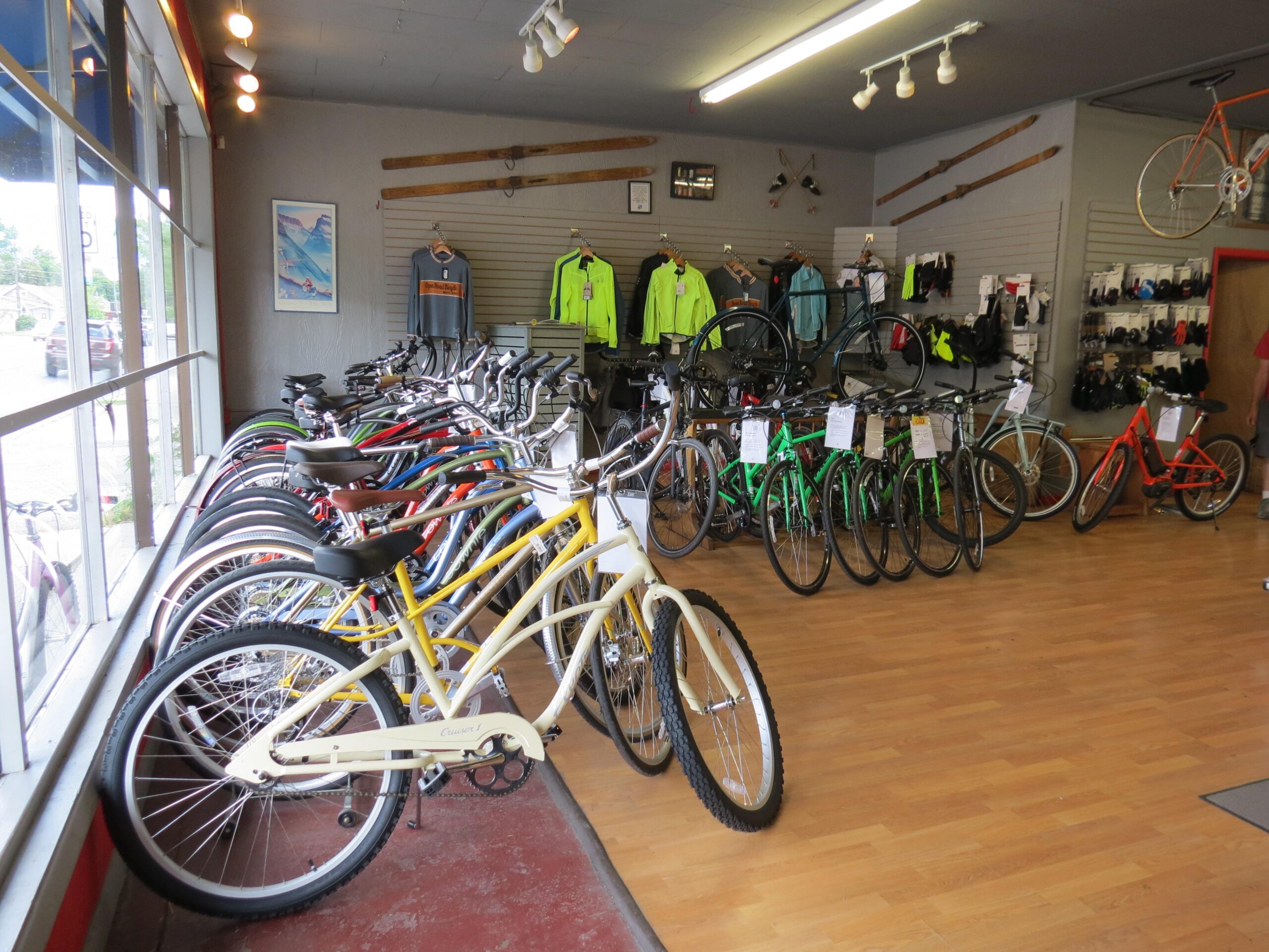 A brightly lit bicycle shop featuring a variety of bicycles in different colors, including yellow, red, green, and white. The shop has wooden flooring and displays sporting apparel and accessories on the walls. Bicycles are arranged in rows, with some hanging from the ceiling. Large windows showcase an outdoor view.