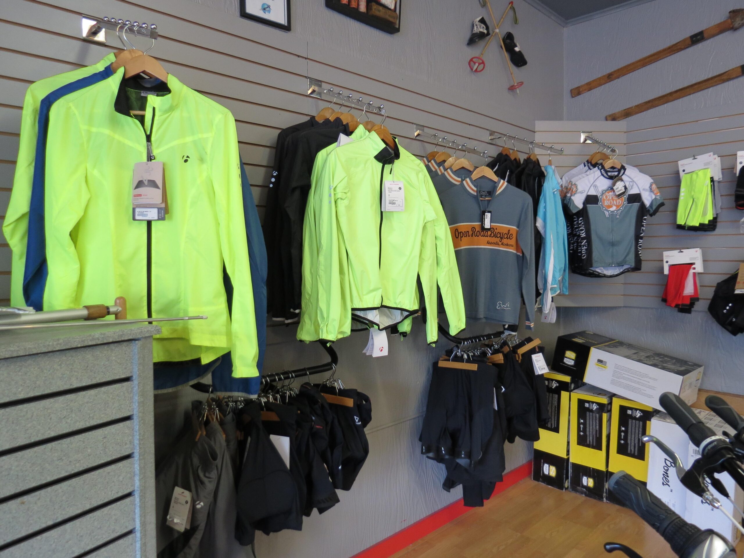 A display of various cycling apparel in a store, featuring bright neon yellow jackets, sleek bike shorts, and jerseys hanging on a wall. The merchandise includes a mix of styles, colors, and sizes, organized on wooden hangers. In the foreground, a counter can be seen with some biking accessories and packaged items near a bicycle.