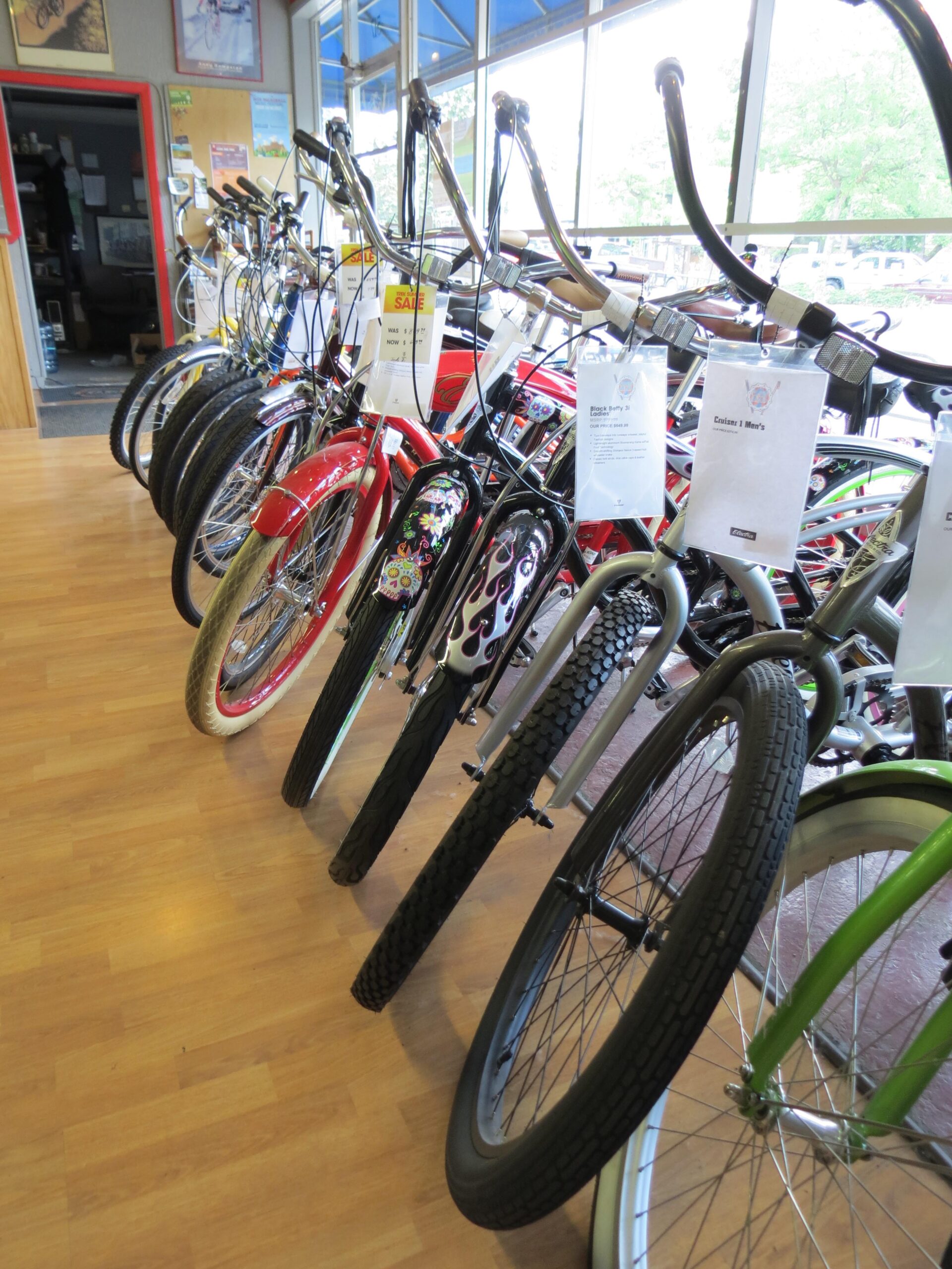 A row of various bicycles displayed in a shop interior, showcasing different styles and colors. The bikes have distinct frames and tires, with some featuring decorative designs. Price tags and information sheets are attached to the handlebars. Natural light comes through large windows in the background, illuminating the wooden floor and creating a welcoming atmosphere.