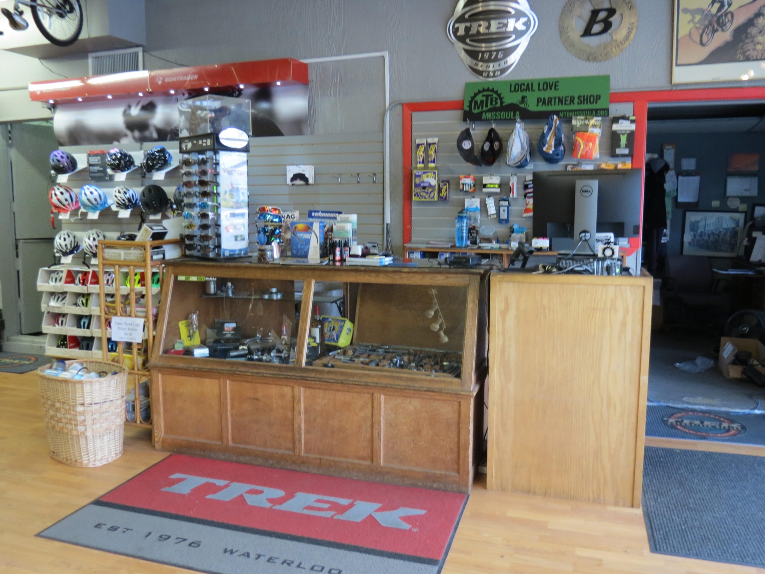 Alt text: Interior of a bicycle shop featuring a wooden display counter with bike tools, helmets hanging on the wall, and a computer checkout area. The store has a wooden floor and a "Trek" logo mat at the entrance.