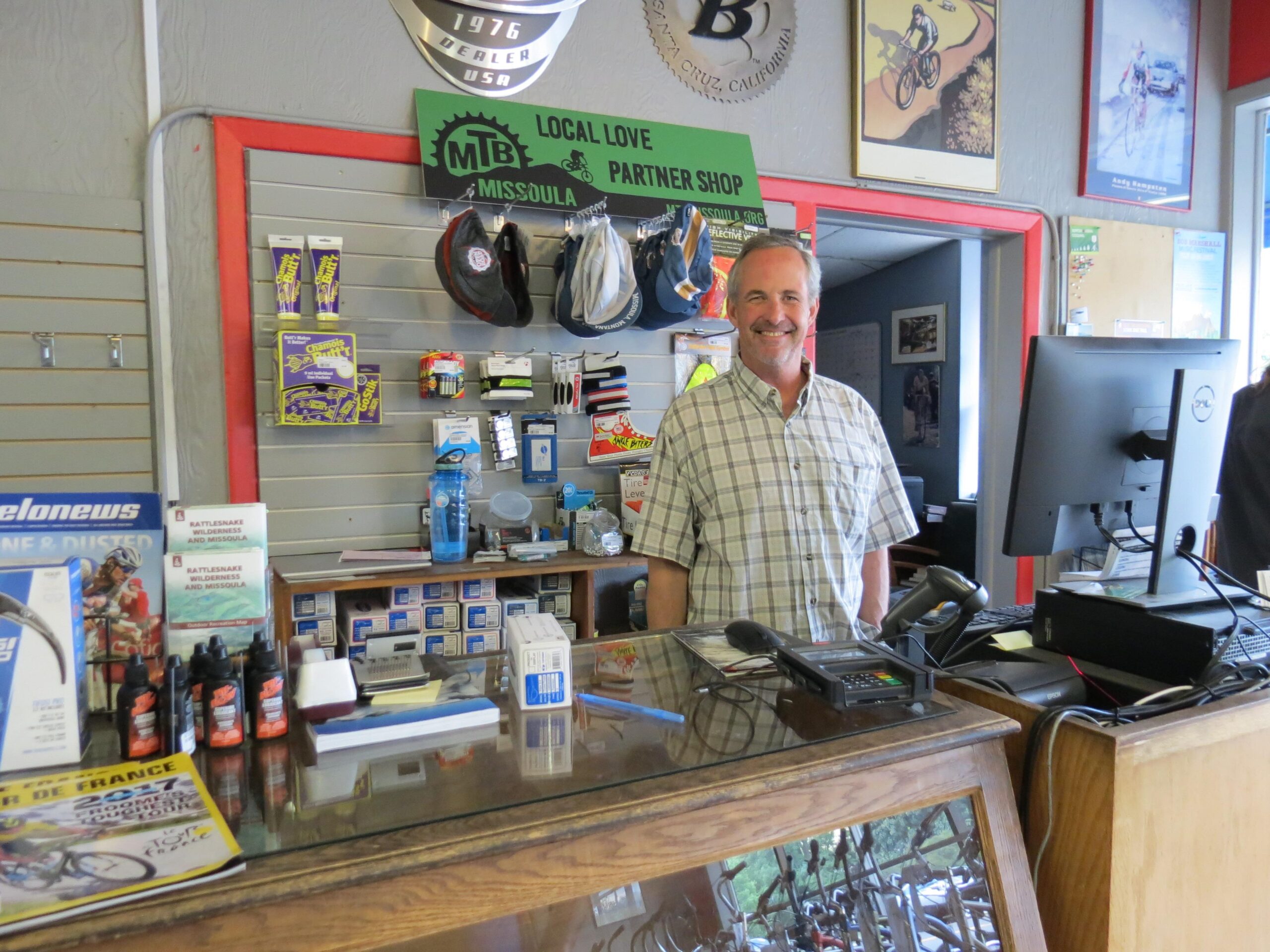 A smiling shopkeeper stands behind a counter in a bicycle shop. The background features shelves stocked with cycling accessories and helmets, along with posters of bicycles. A computer and various items are displayed on the counter, showcasing a welcoming atmosphere for customers. A sign indicating the shop is a local partner is visible on the wall.