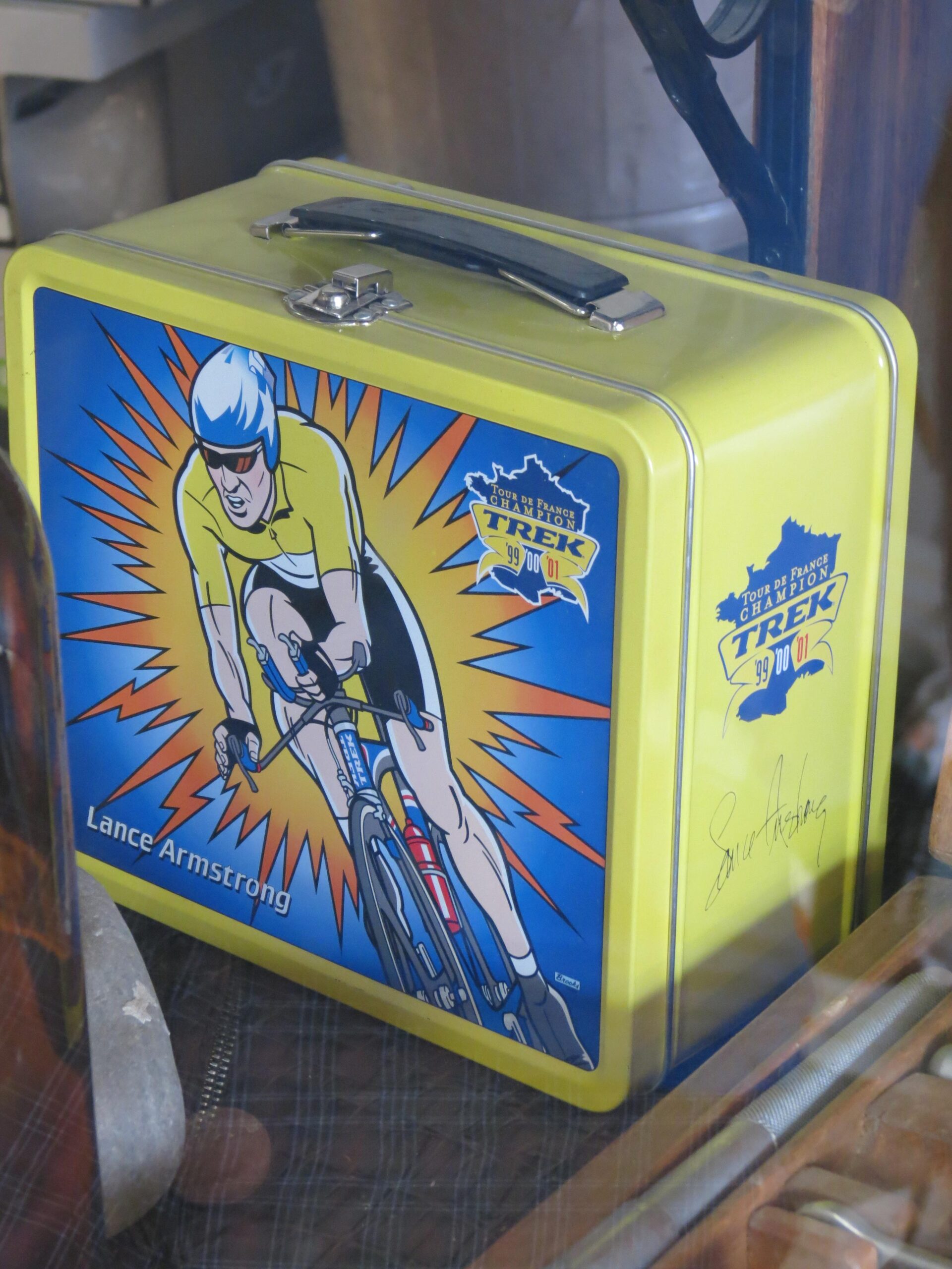 A vintage yellow lunchbox featuring an illustration of a cyclist in a racing uniform, with bold colors and dynamic lines. The design includes references to the Tour de France and the Trek brand, along with the name "Lance Armstrong" prominently displayed. The lunchbox is presented in a glass case, reflecting a nostalgic appeal.