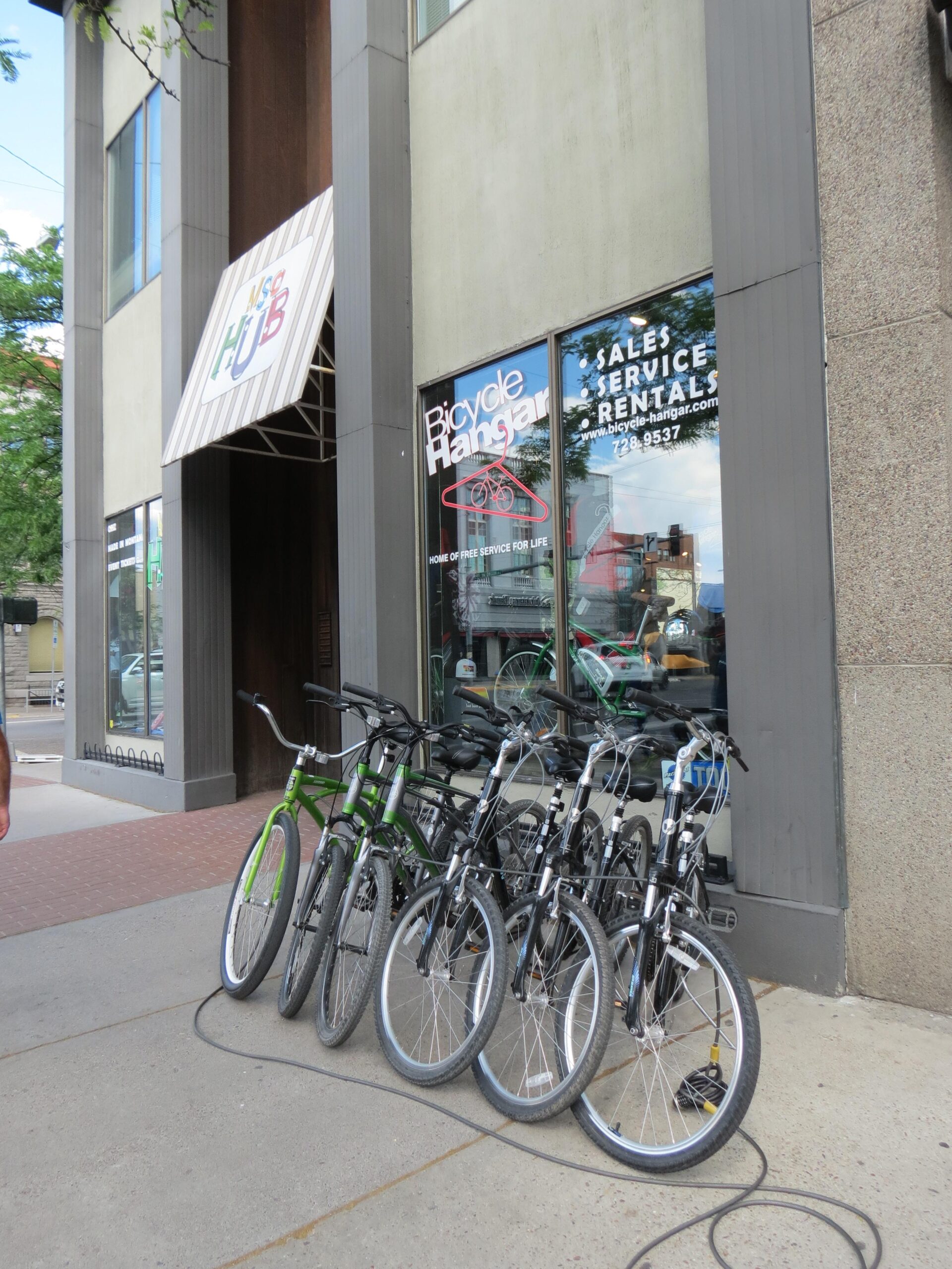 Row of bicycles parked outside a store with the sign "Bicycle Hangar" displayed in the window, offering sales, service, and rentals. The storefront also features an awning with the logo of "The Hub."