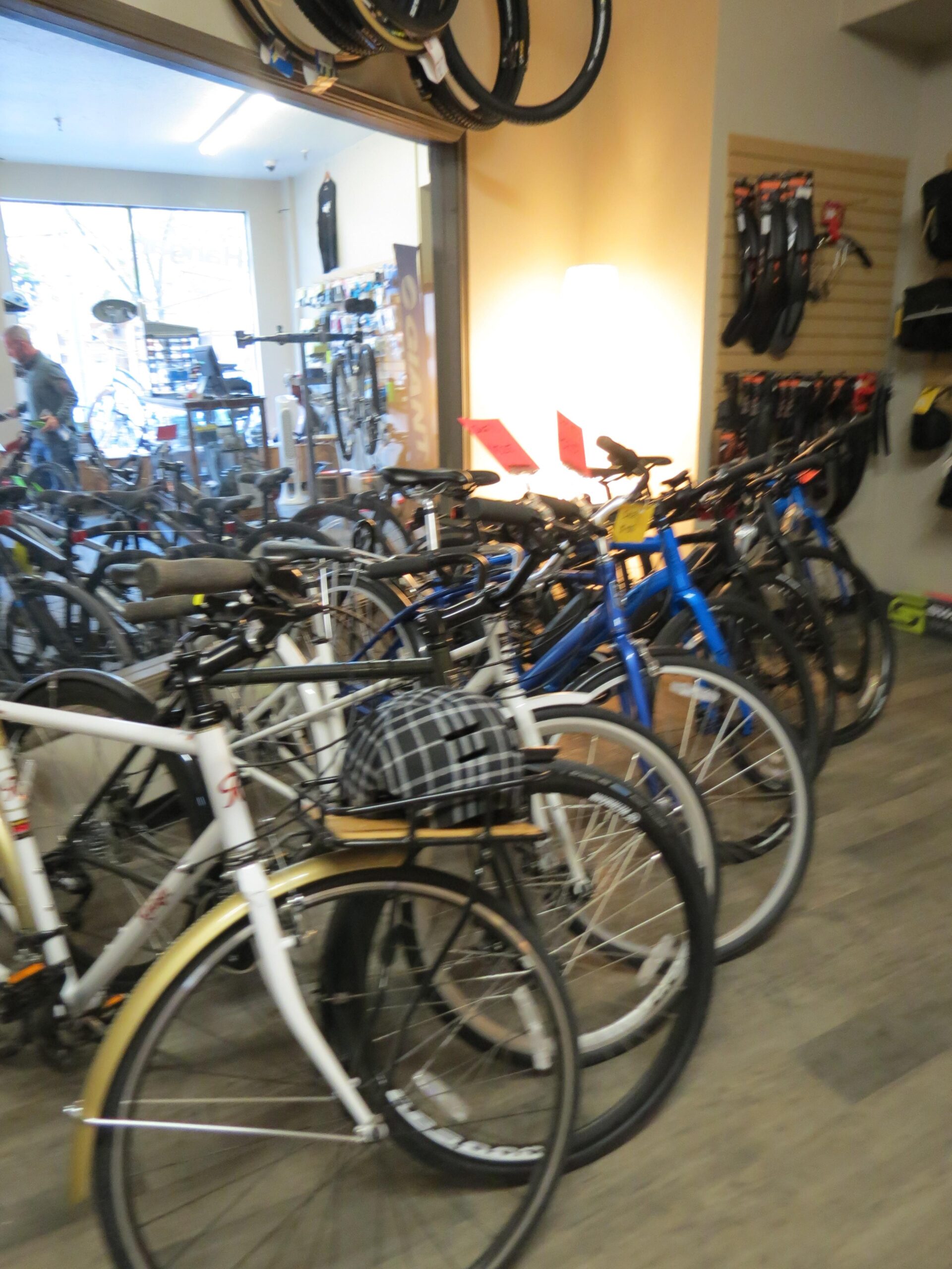 A busy bicycle shop featuring a variety of bikes for sale, including several in different colors. In the foreground, a white bike with a plaid helmet on its rack is visible. The shop is well-lit, with bike accessories displayed on the walls and a sales counter in the background.