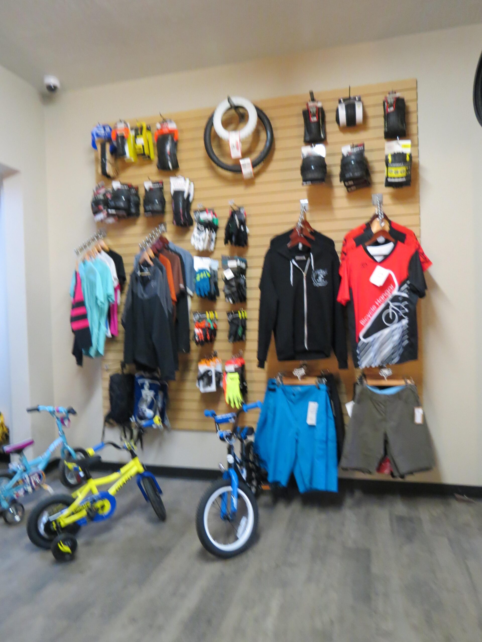 Interior view of a bike shop displaying various cycling apparel and accessories. The wall features a variety of gloves, shirts, and shorts alongside bike tires. In the foreground, there are two children's bicycles, one blue and one yellow. The flooring is wood-like, and the environment appears bright and organized.