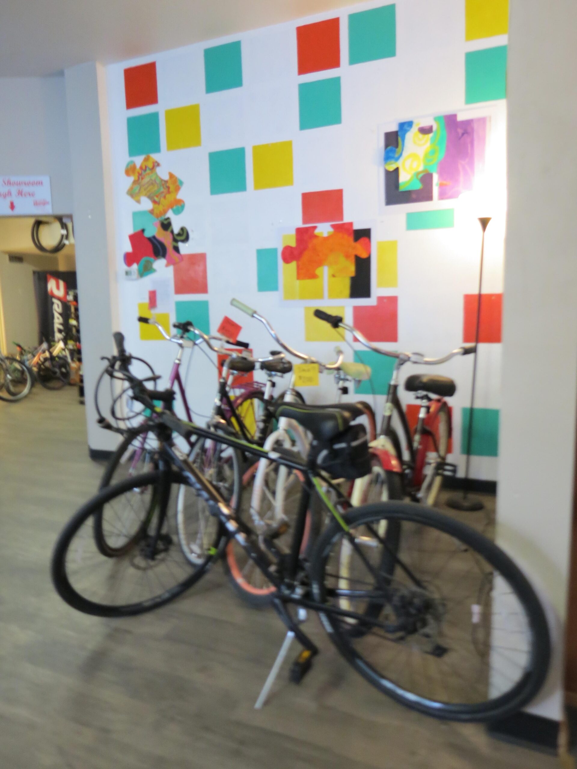 A group of bicycles parked in a brightly colored room with a decorative wall featuring squares and puzzle piece art. The scene also includes a standing lamp.