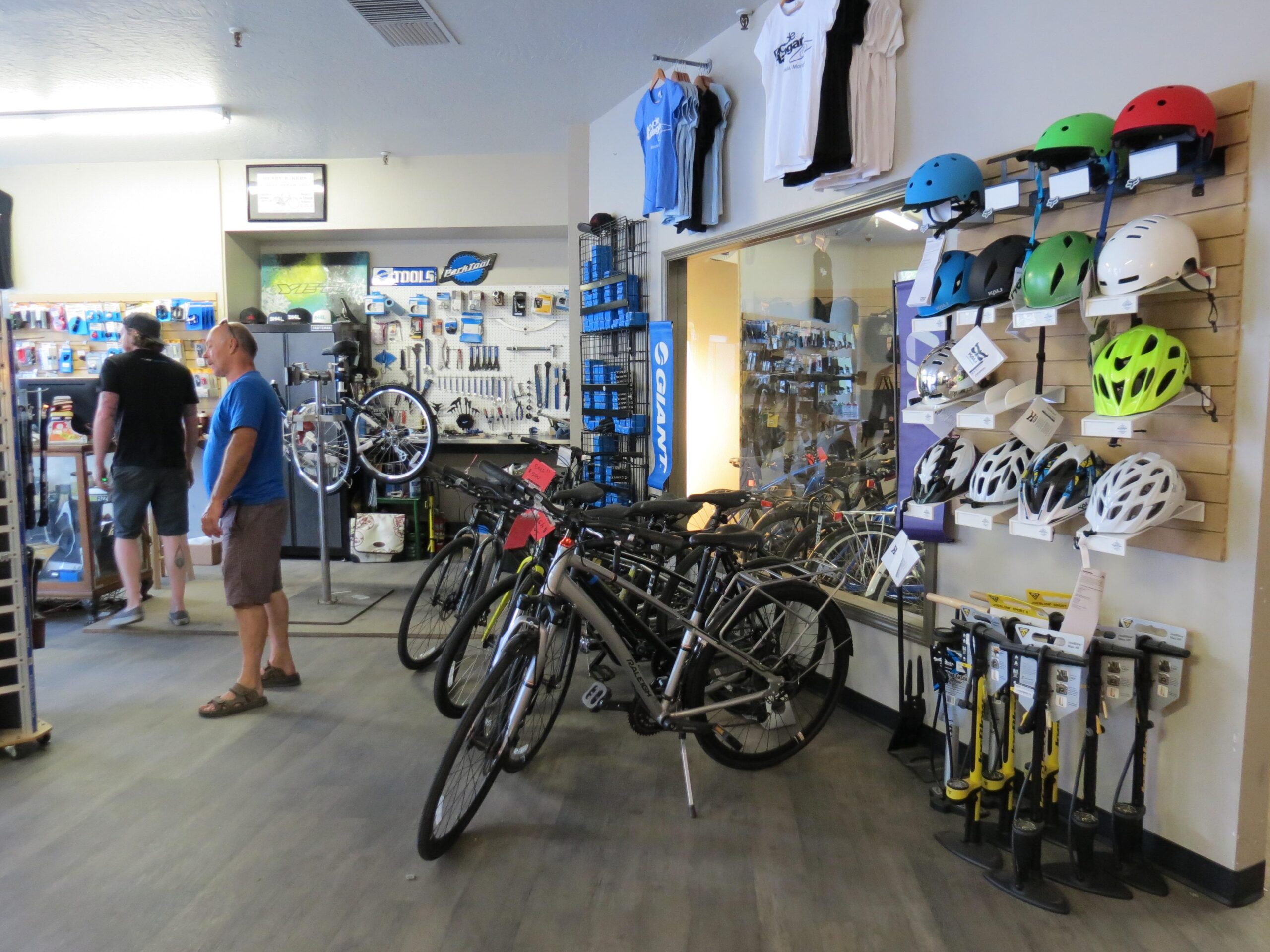 A view of a bicycle shop interior featuring various bicycles and cycling equipment. There are several helmets displayed on the wall in different colors, along with tools and accessories organized on shelves. Two customers are browsing, one standing near the entrance and another at the back of the shop. The atmosphere is bright and inviting, with merchandise neatly arranged.