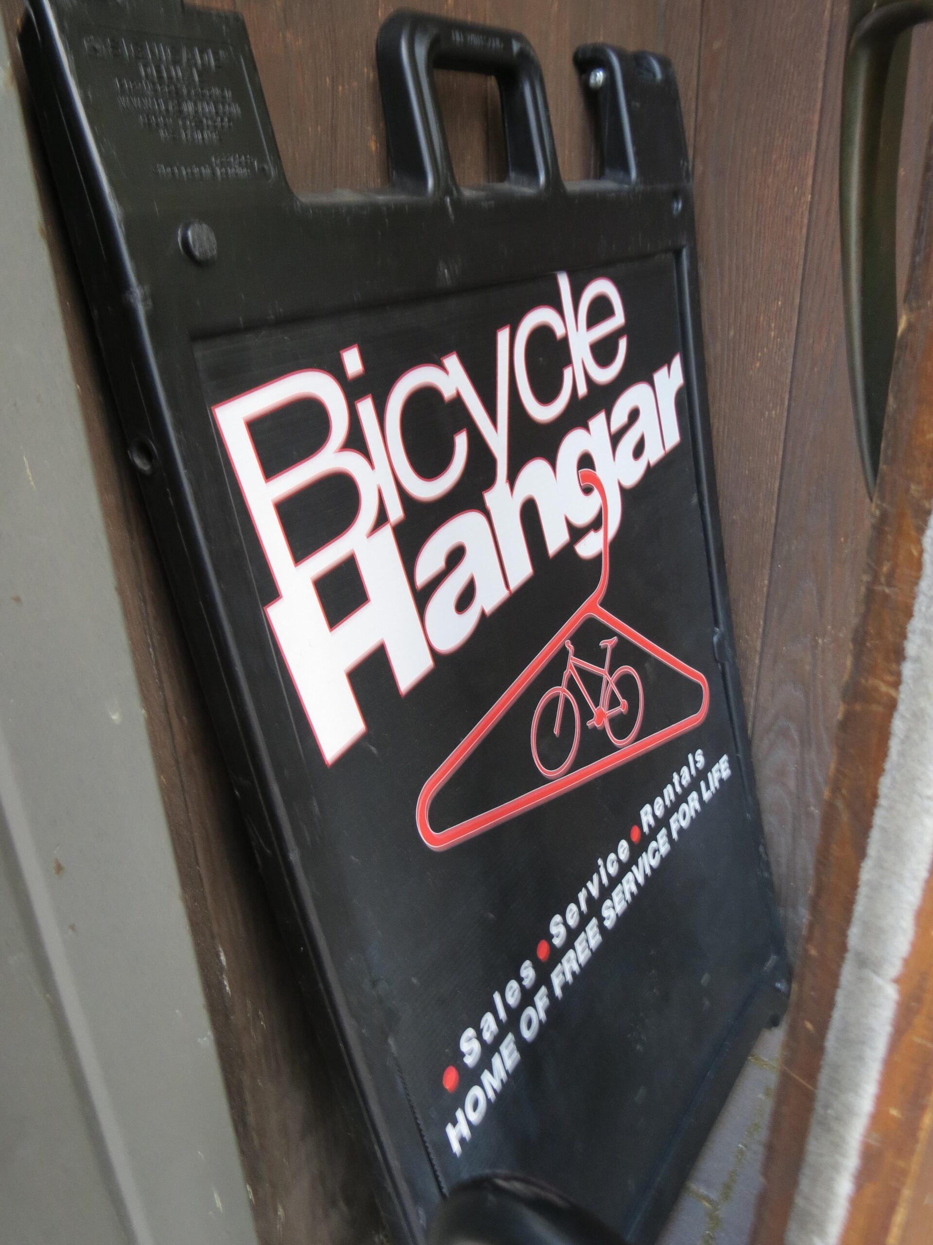 A black roadside sign for "Bicycle Hangar," featuring white and red text indicating sales, service, and rentals. The sign highlights "Home of Free Service for Life," with an illustration of a bicycle. The background shows a wooden surface.