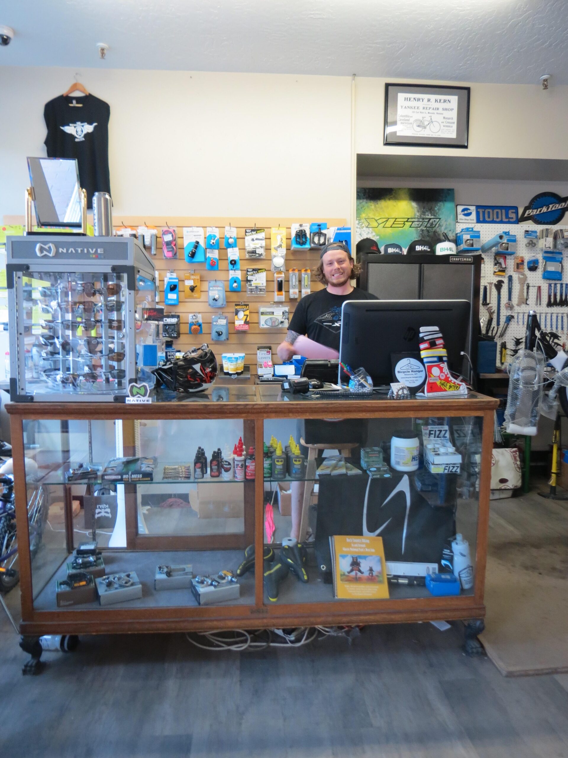 A bike repair shop interior featuring a smiling employee behind a wooden counter. The wall is adorned with various biking accessories and tools, and a display case showcases additional items. The space is well-organized, with numerous biking products visible in the background.