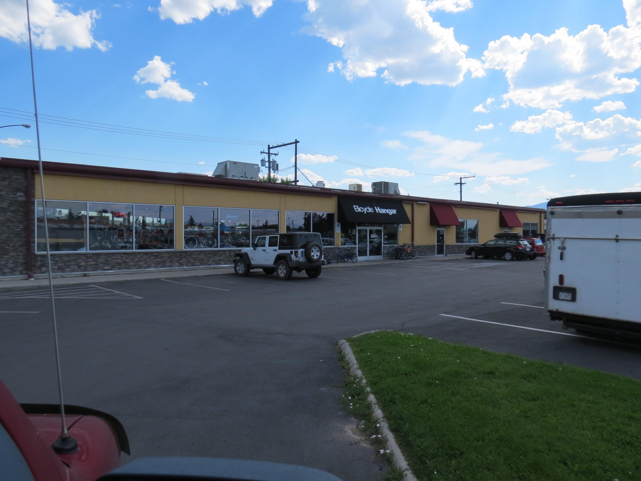 A view of a bicycle shop named "Bicycle Hangar" featuring large windows displaying bikes. The store is located in a parking lot with several vehicles, including a white SUV and a trailer, and a green grassy area in the foreground. The sky is partly cloudy with blue hues.