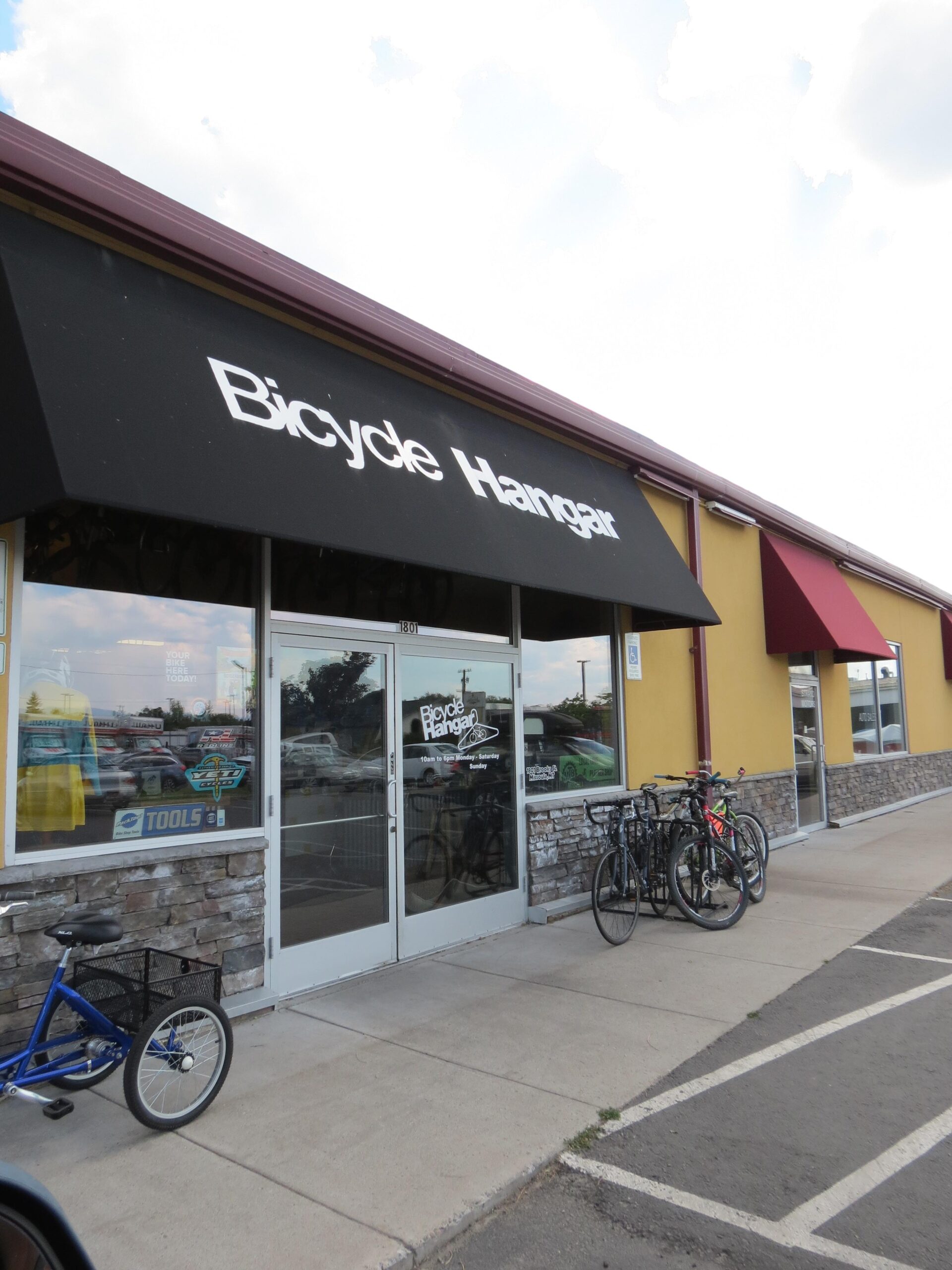 Exterior view of Bicycle Hangar store, featuring a prominent black awning with the store name displayed. The entrance has glass doors, and several bicycles are parked outside, including a blue tricycle and various other bikes. The building has a stone veneer and is adjacent to a yellow-painted structure. The sky is partly cloudy.