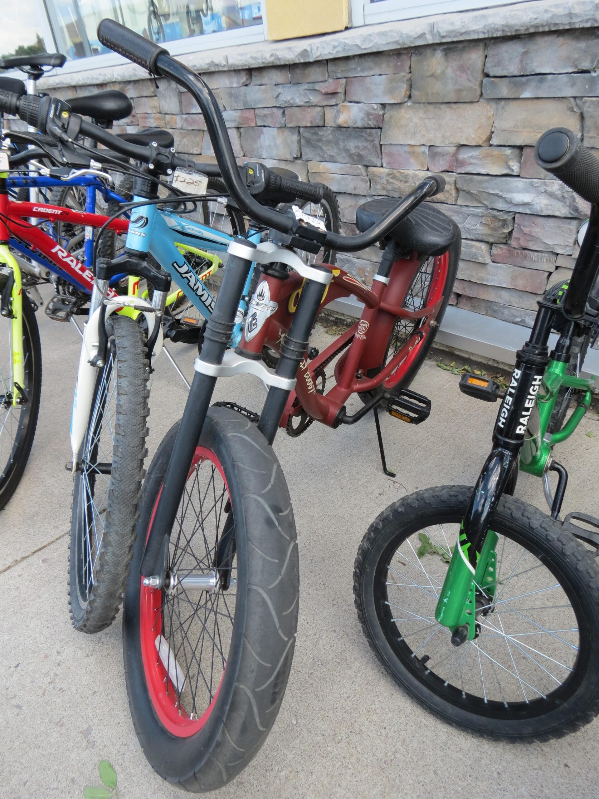 A close-up view of several bicycles parked next to a stone wall. The image features a variety of bike styles, including a BMX bike with a thick, red-rimmed tire, a mountain bike with a blue frame, and a smaller green bike. The bicycles are lined up closely together, showcasing different colors and designs.