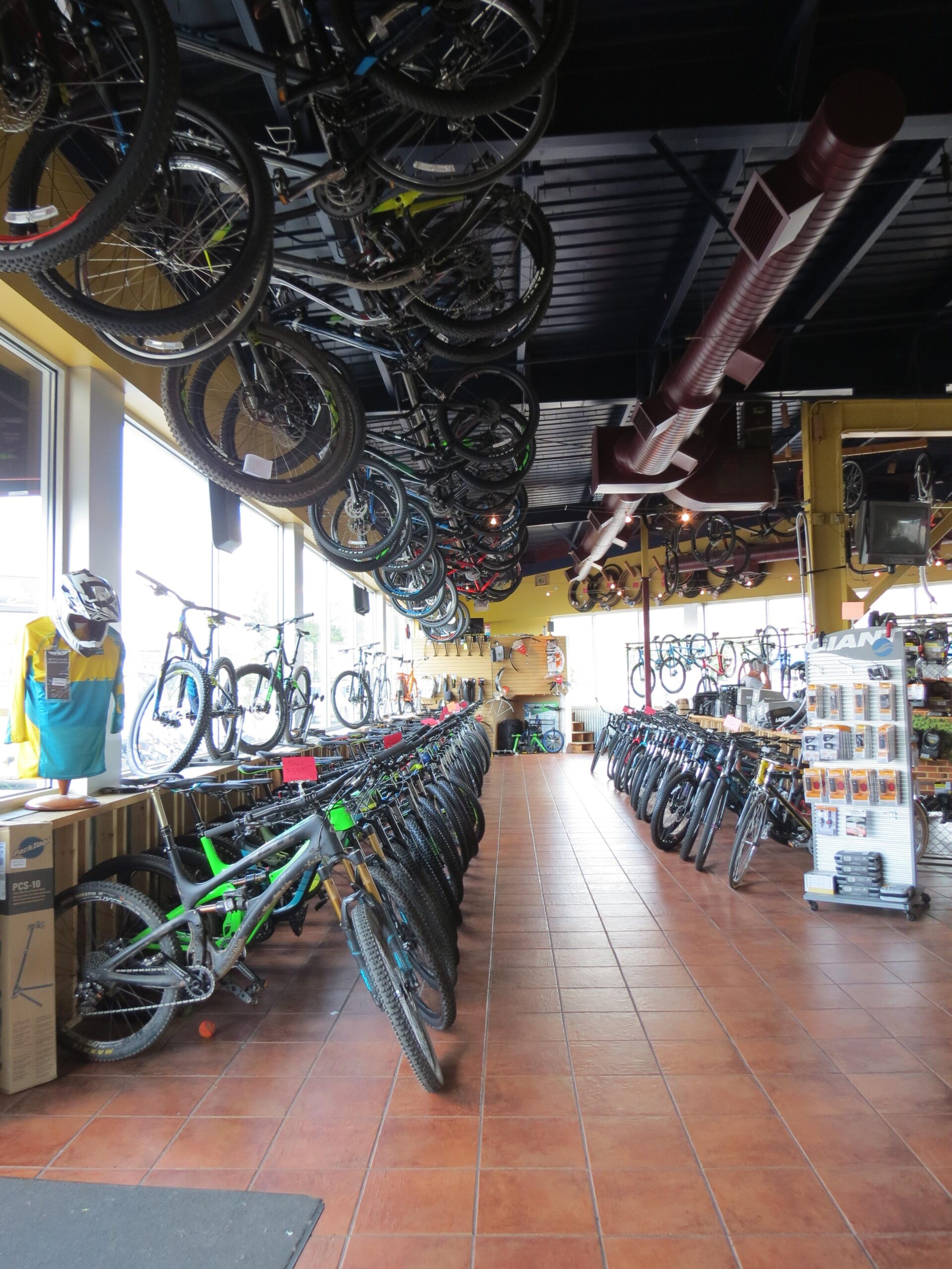 A bike shop interior featuring a row of mountain bikes displayed prominently on either side, with additional bikes hanging from the ceiling. The shop has a warm atmosphere, highlighted by natural light coming through large windows. Merchandise and accessories are also visible along the walls and in display units.