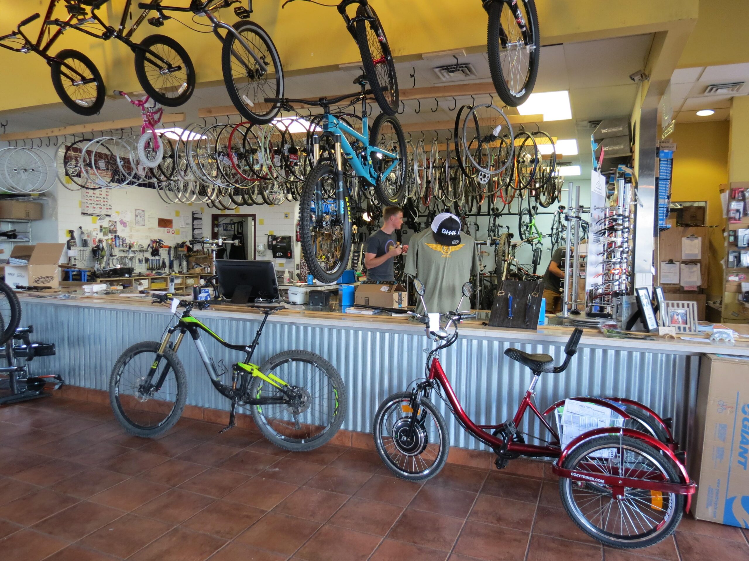 A bustling bicycle shop interior featuring various bikes hanging from the ceiling and displayed on the floor. Two staff members are engaged in work at the counter, with tools and equipment visible in the background. The shop has a bright yellow and gray color scheme, creating a lively atmosphere.