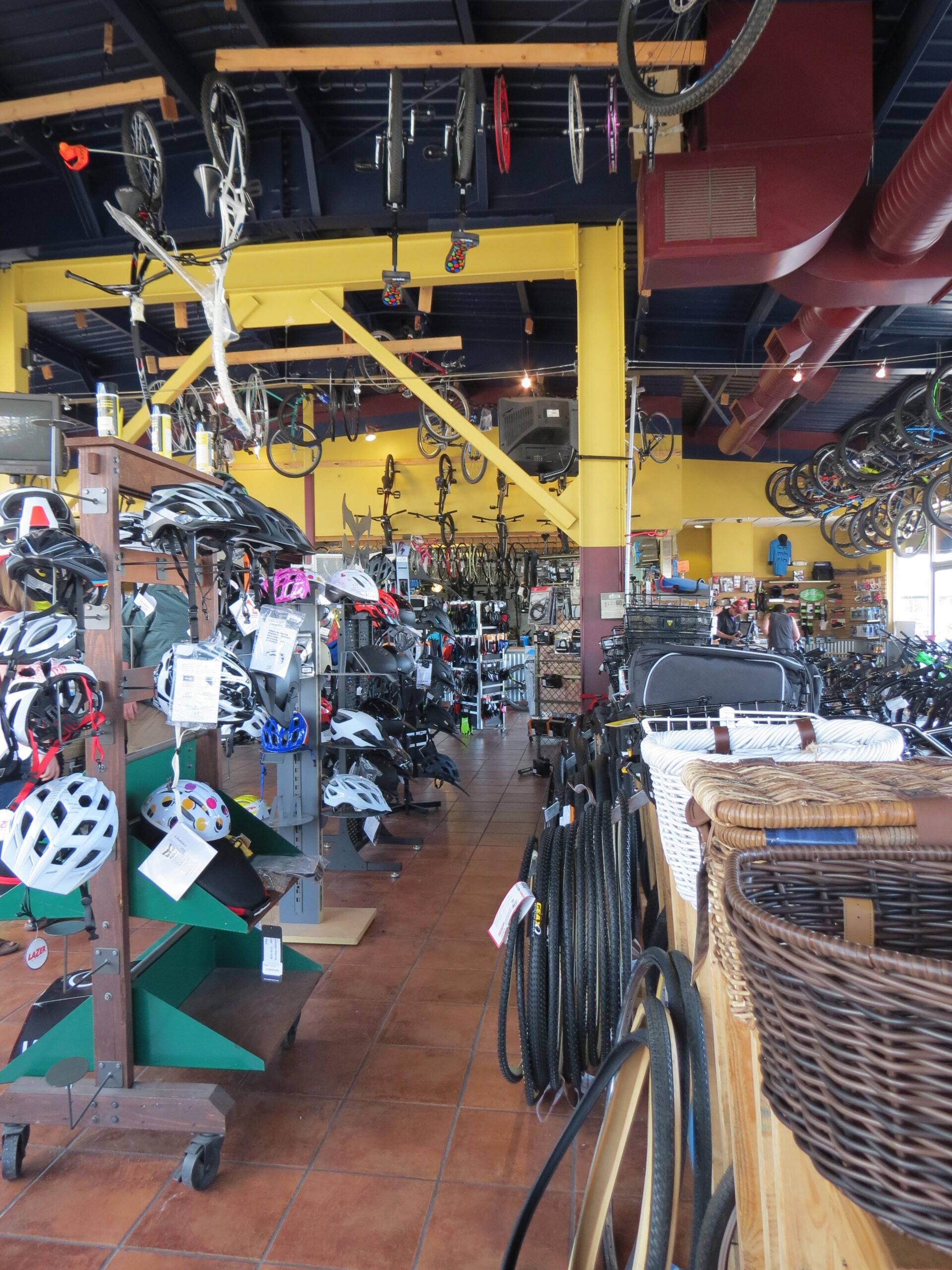 Interior view of a bicycle store displaying a variety of bikes hanging from the ceiling and shelves stocked with helmets, bike tires, and other cycling accessories. The vibrant yellow and blue decor adds to the lively atmosphere of the shop.