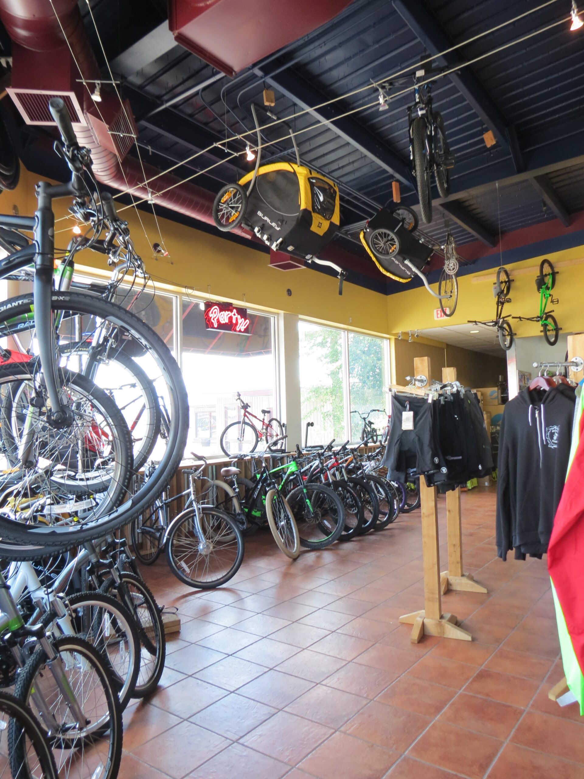 Interior of a bike shop featuring various bicycles displayed on the floor and hanging from the ceiling. In the foreground, several bikes are lined up against the walls, while a yellow bike trailer is suspended from the ceiling. The shop has a bright, open feel with a tiled floor and a few clothing items hung on wooden displays. Natural light is coming in through large front windows.