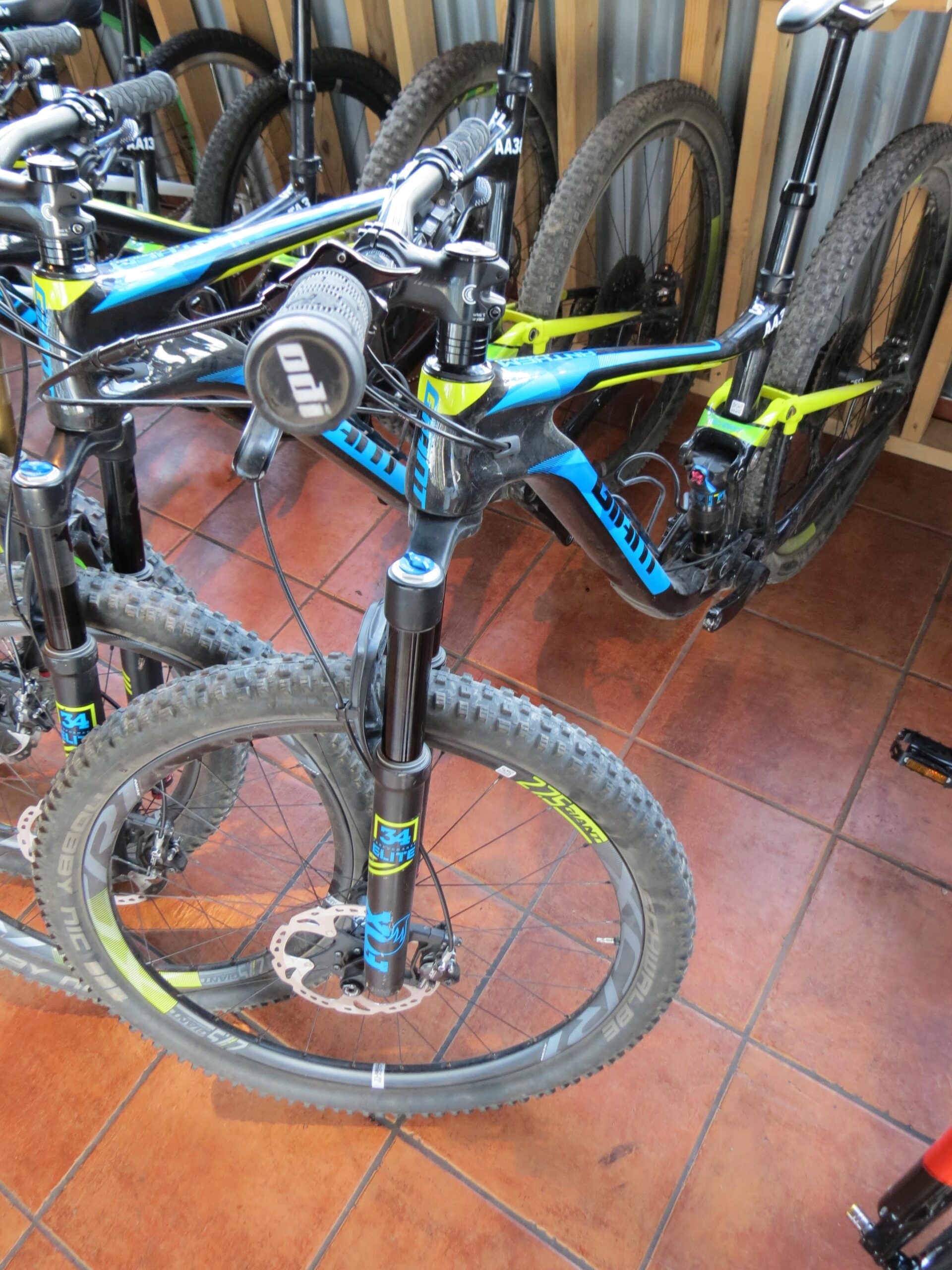 A close-up view of a mountain bike with a blue and yellow frame, displaying the handlebars and front wheel. The bike is positioned among other similar bikes in a storage area with wooden walls and a tiled floor. The photo captures details like the tires, brake components, and handlebars, emphasizing its design and features.