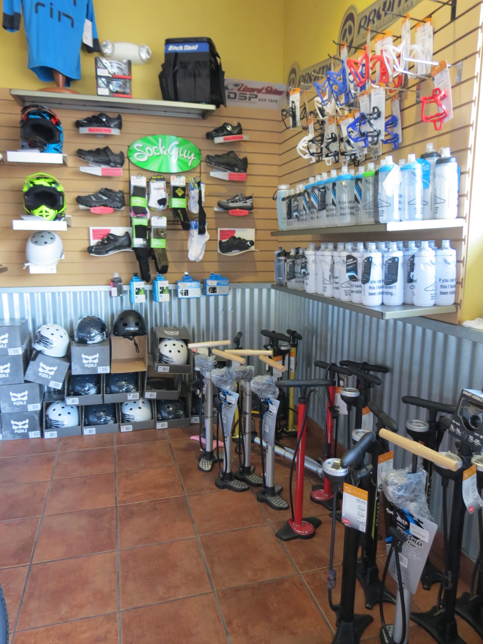 A brightly lit interior of a sports equipment store showcasing a variety of cycling gear. The wall features colorful helmets, cycling shoes, socks, and water bottles, with merchandise labeled on price tags. A selection of bicycle pumps is displayed prominently on the floor, alongside stacked boxes of helmets. The store's design blends functional display with an appealing layout for customers.