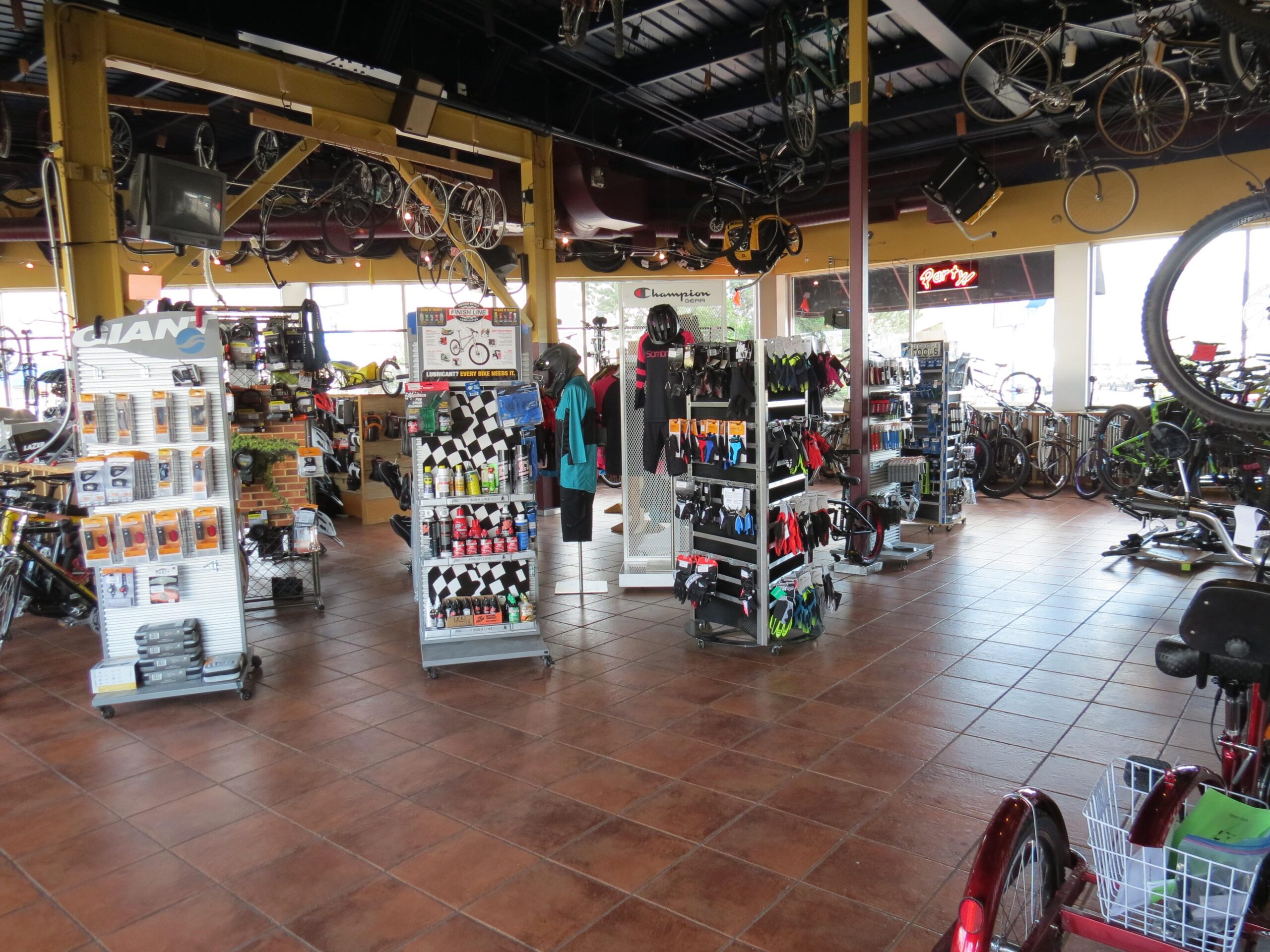 A spacious bicycle shop interior featuring a variety of bicycles on display, accessories organized on shelves, and bike gear hanging from the ceiling. Brightly lit with large windows allowing natural light, the store has tiled flooring and a welcoming atmosphere. Several racks showcase items like helmets, gloves, and maintenance products.
