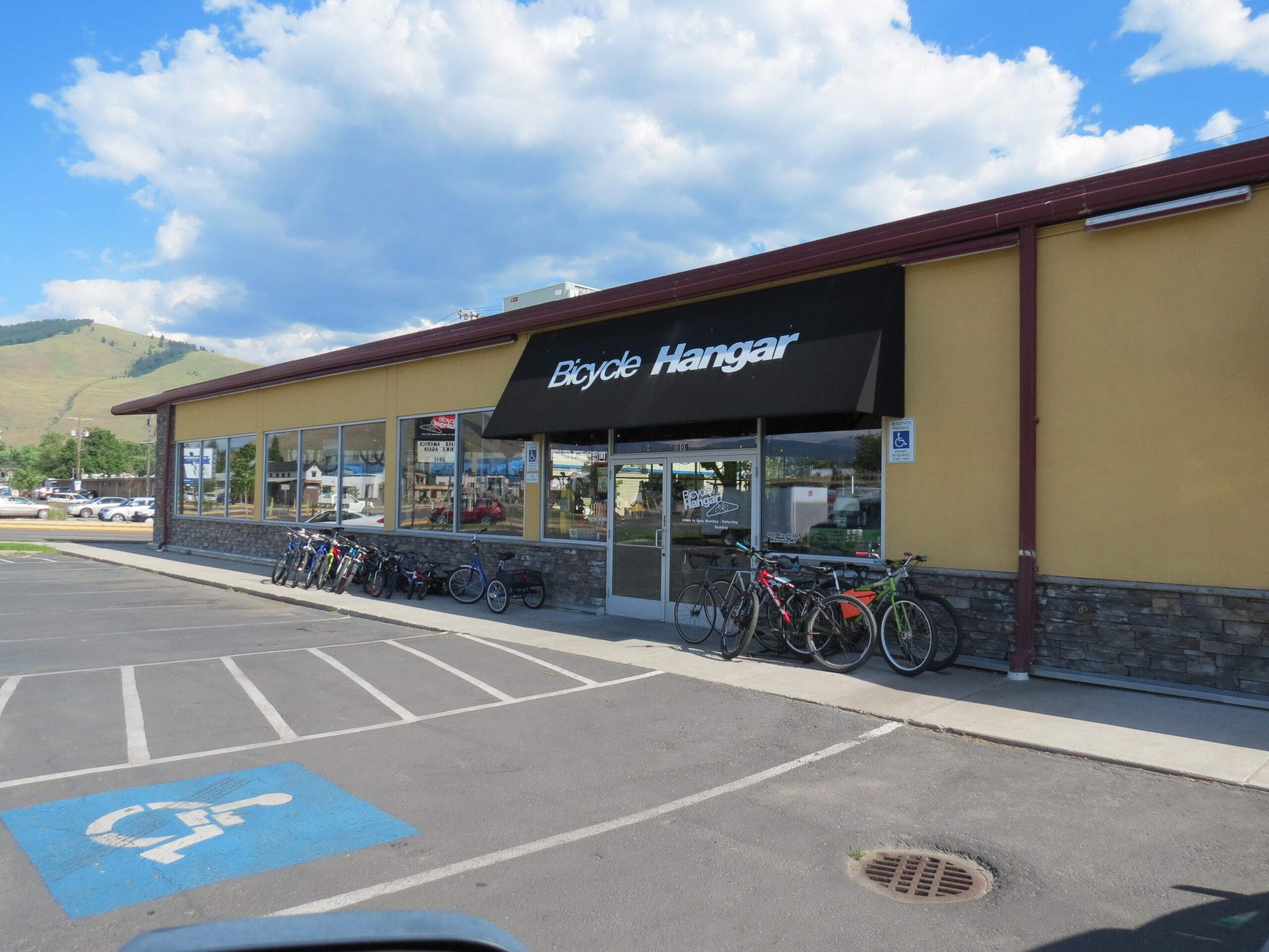A bicycle shop called "Bicycle Hangar," featuring a large window front and an awning. Several bicycles are parked outside in a designated bike area, with a handicap parking space visible in the foreground. The backdrop includes rolling hills under a partly cloudy sky.