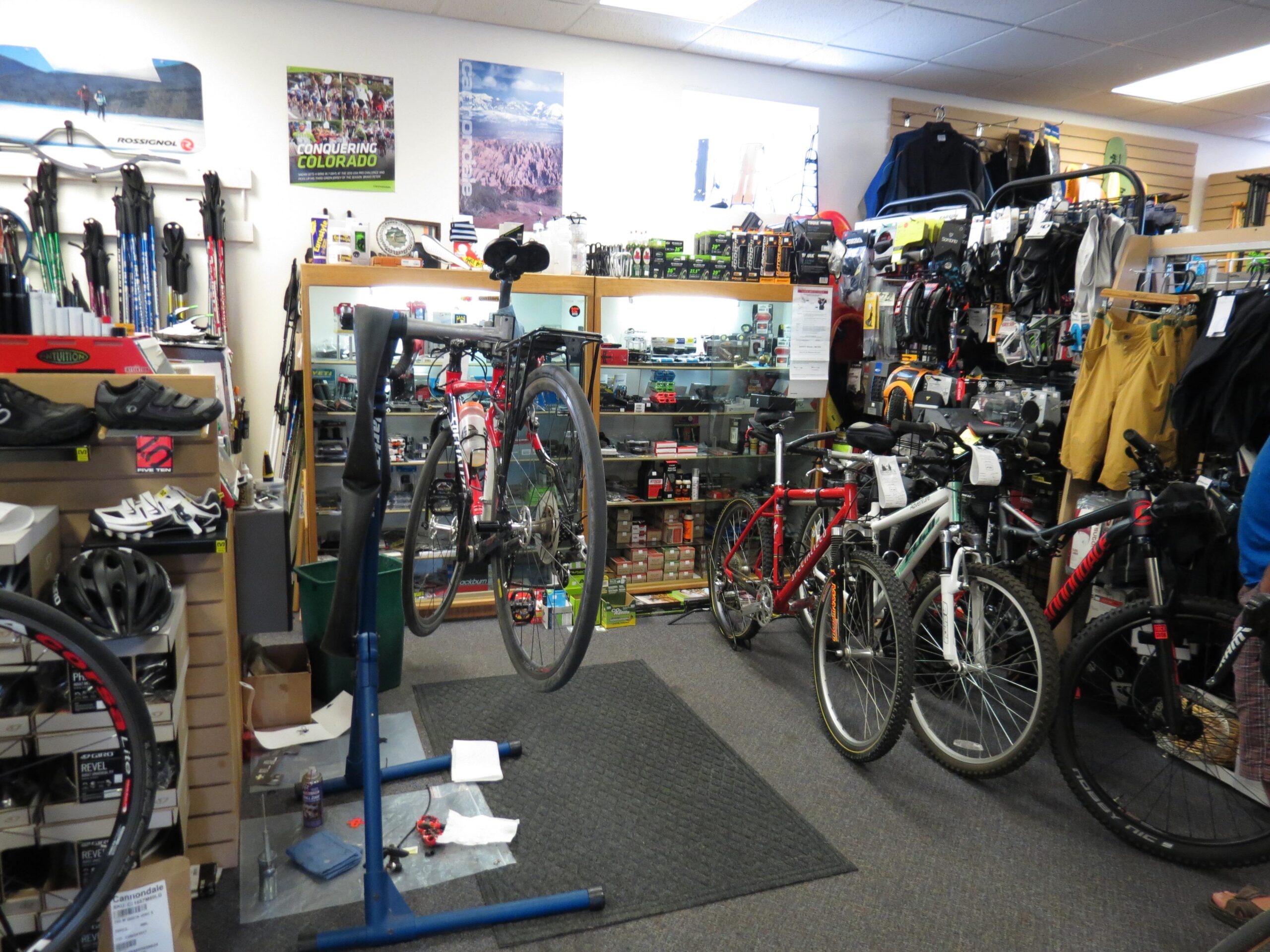 A busy bicycle shop interior displaying various bikes, helmets, and cycling gear. A bike is positioned on a repair stand in the center, while additional bicycles are parked nearby. Shelves feature an assortment of cycling accessories and merchandise, with promotional posters on the walls.