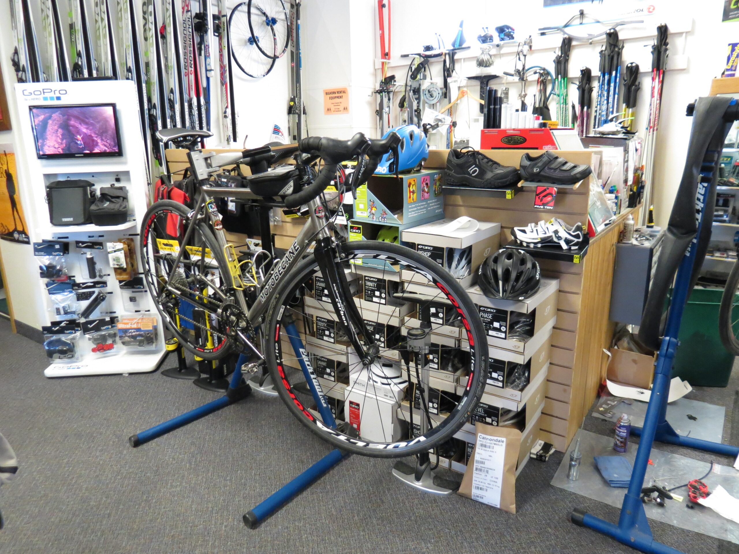 Alt text: A bicycle is displayed on a repair stand in a storefront filled with cycling and outdoor gear. Various shoes, accessories, and a GoPro display can be seen in the background, along with ski equipment mounted on the wall.