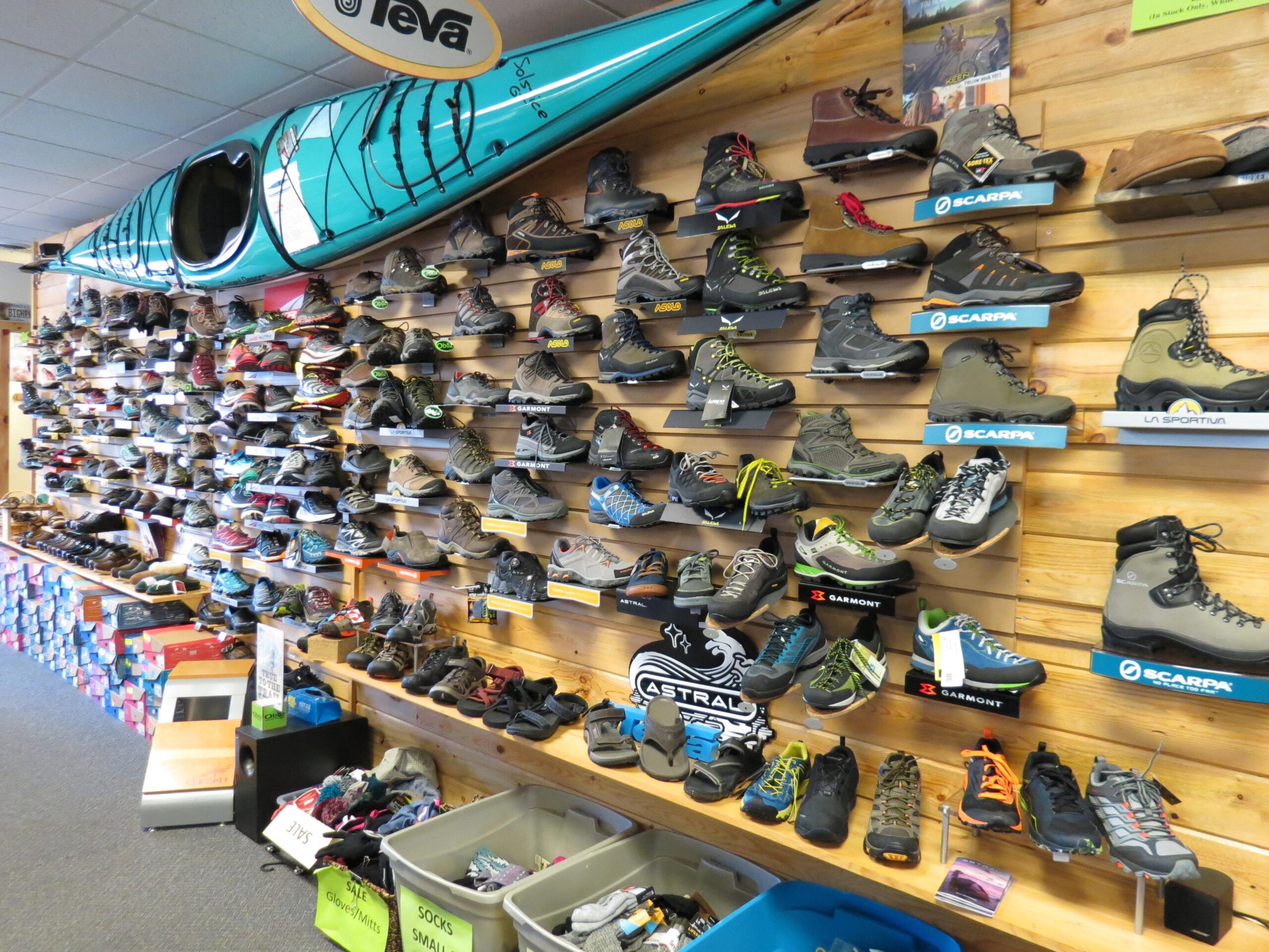 A retail display showcasing a variety of hiking and outdoor footwear on a wooden wall. The shoes are arranged on shelves of different heights, with brands prominently labeled. A bright turquoise kayak hangs overhead, adding to the outdoor theme. In the foreground, there are bins with socks and mittens, and a stack of shoeboxes is visible. The space appears well-organized and inviting for outdoor enthusiasts.