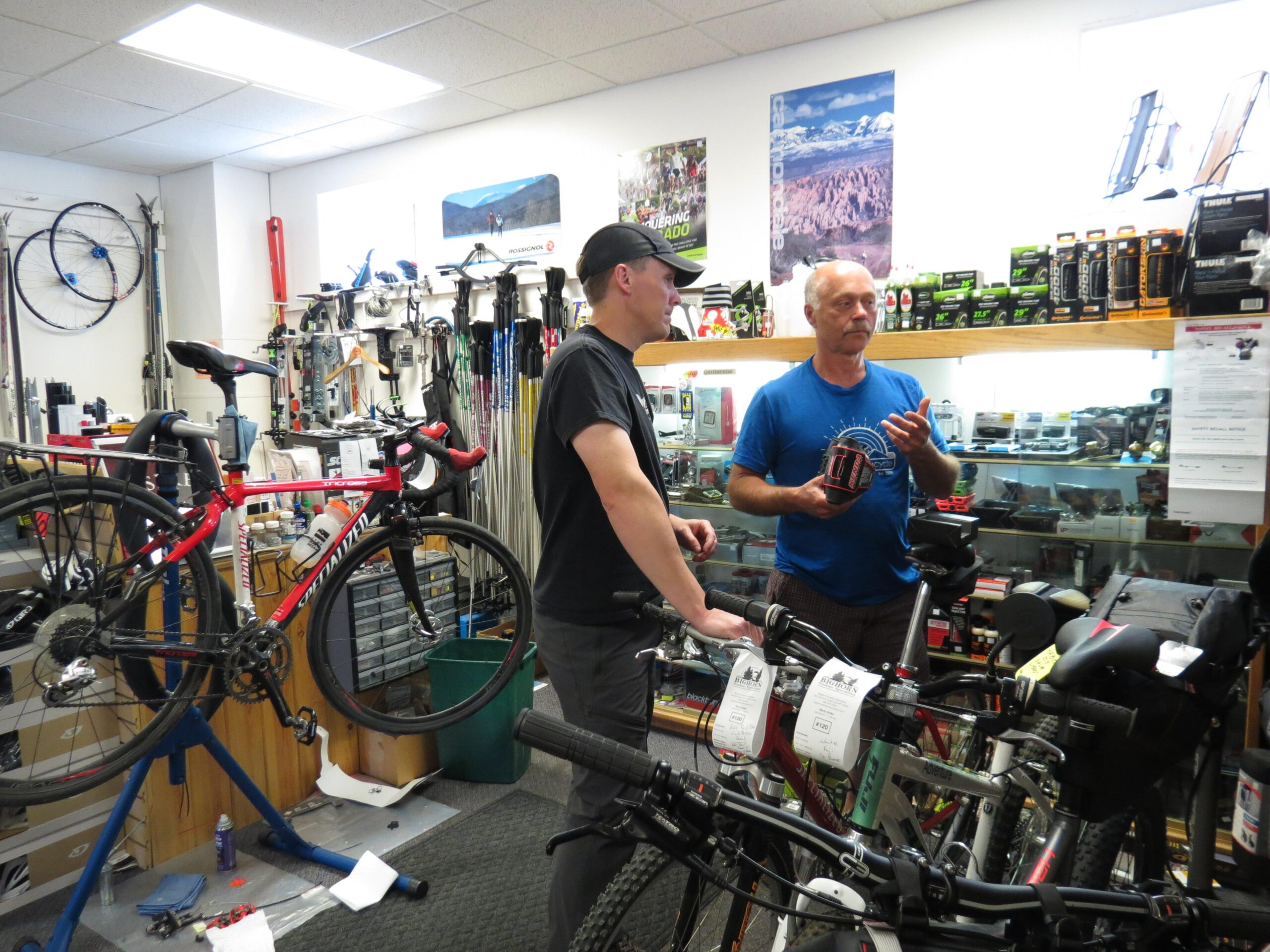 Two men engage in conversation in a bicycle shop, surrounded by various bikes and accessories. One man is holding a product while the other listens attentively, with bicycles prominently displayed in the background. The shop is well-organized, featuring tools and equipment for cycling, contributing to a vibrant retail atmosphere.