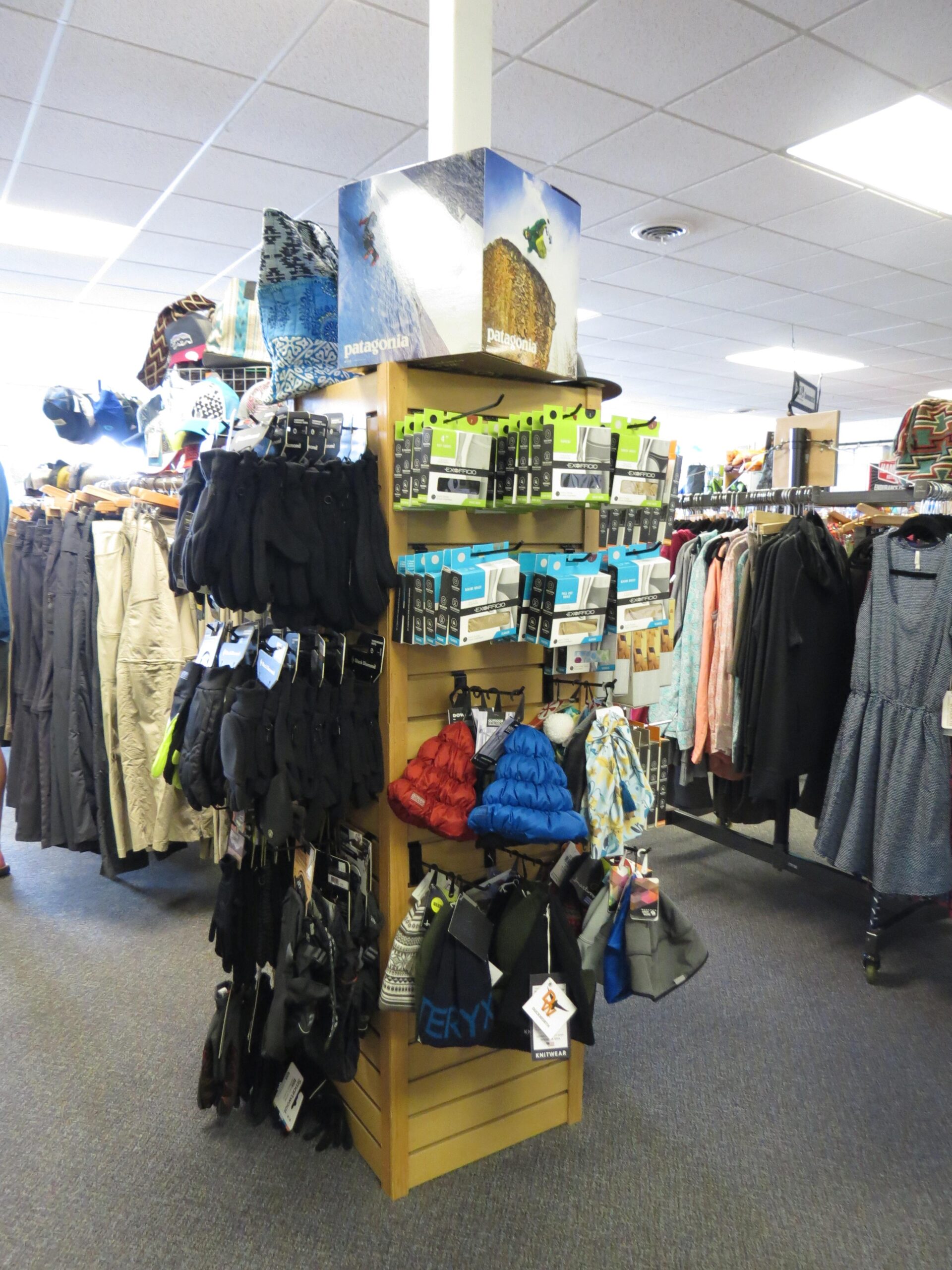 A retail display in a clothing store featuring outdoor apparel and accessories. The display includes various types of gloves, beanies, and jackets, with shelves stacked with products from brands like Patagonia and Mervin. The background shows additional clothing racks and a well-lit store interior.