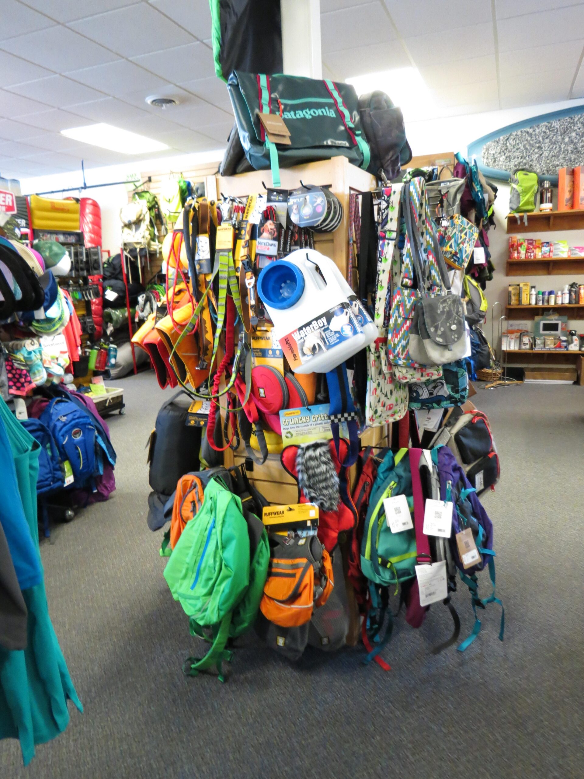 A display of various outdoor bags and backpacks in a retail store, featuring colorful options from brands like Patagonia and others. The arrangement includes multiple styles hanging from a central display, surrounded by additional outdoor gear in the background. The store's interior has bright lighting and a welcoming atmosphere.