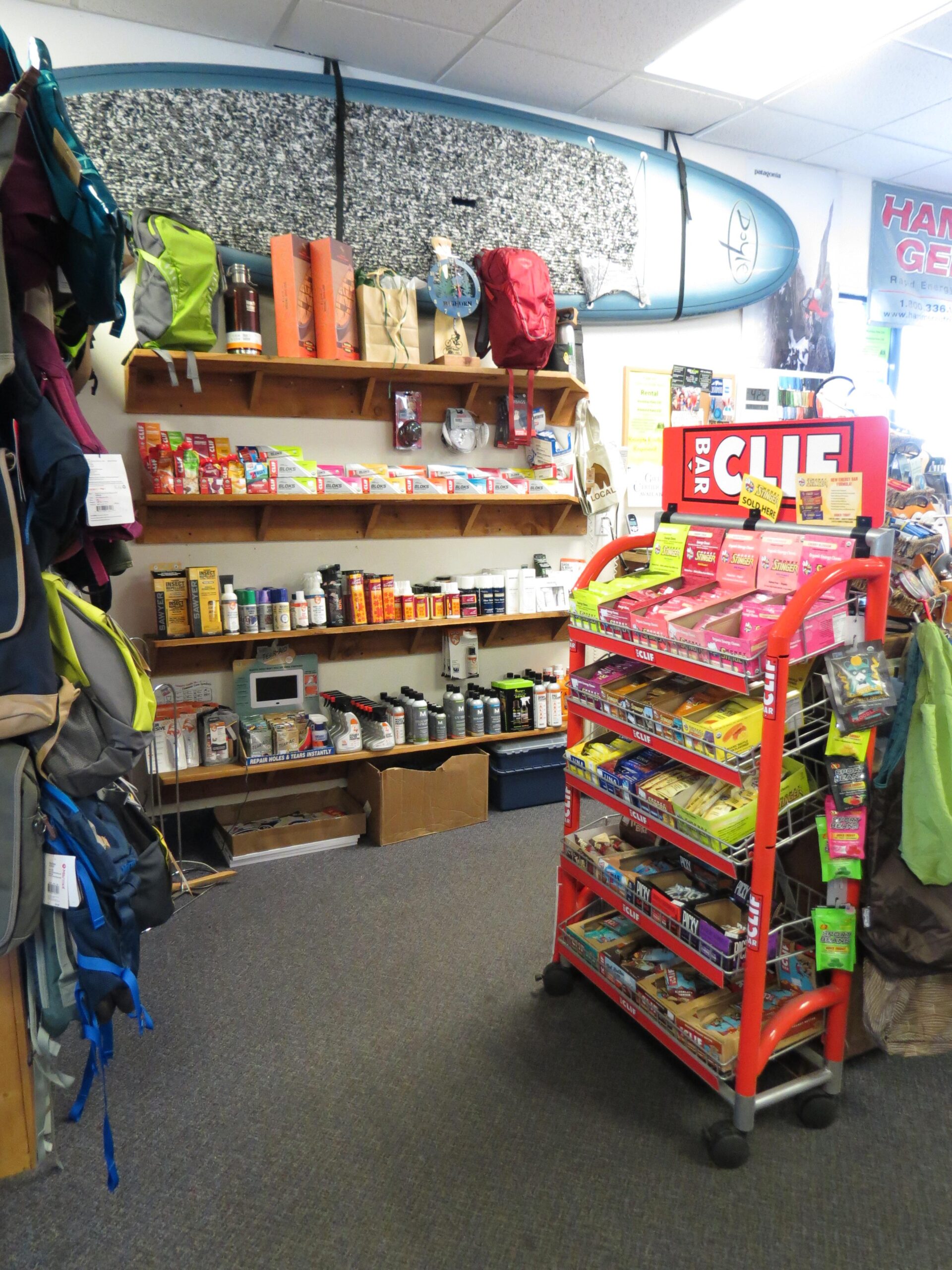 A cozy retail space featuring outdoor gear and snacks. The wall displays a surfboard above wooden shelves stocked with various camping supplies and food items. A colorful display cart holds energy bars and snacks, while several backpacks and bags hang on the side. The shop has a welcoming atmosphere with a clean, organized layout.
