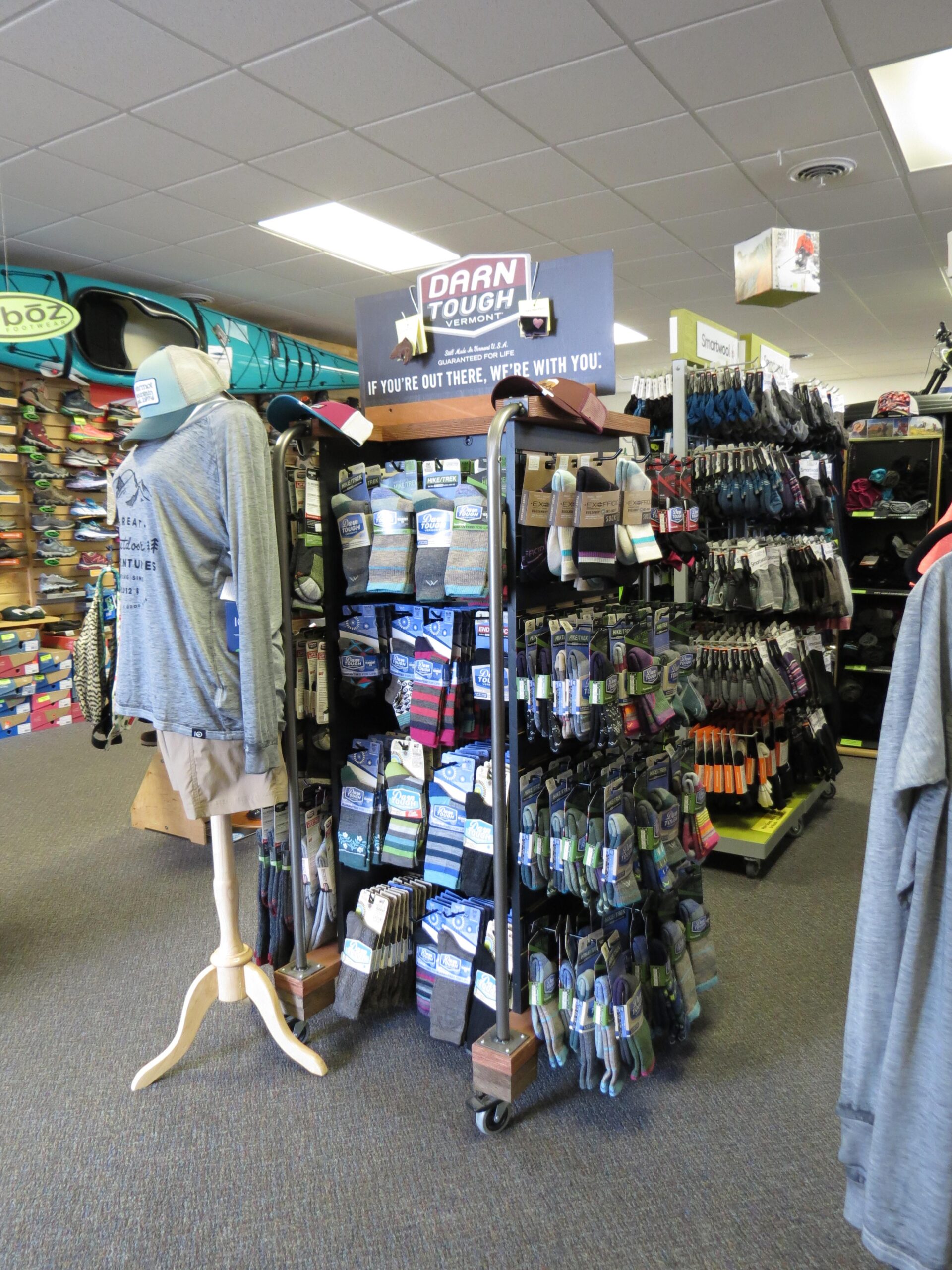 A retail space showcasing outdoor apparel and gear, featuring a display for Darn Tough socks. The area includes a mannequin dressed in a gray long-sleeve shirt and shorts, surrounded by various sock styles on racks. A kayak is visible in the background, and the store is well-lit with a clean, organized layout.
