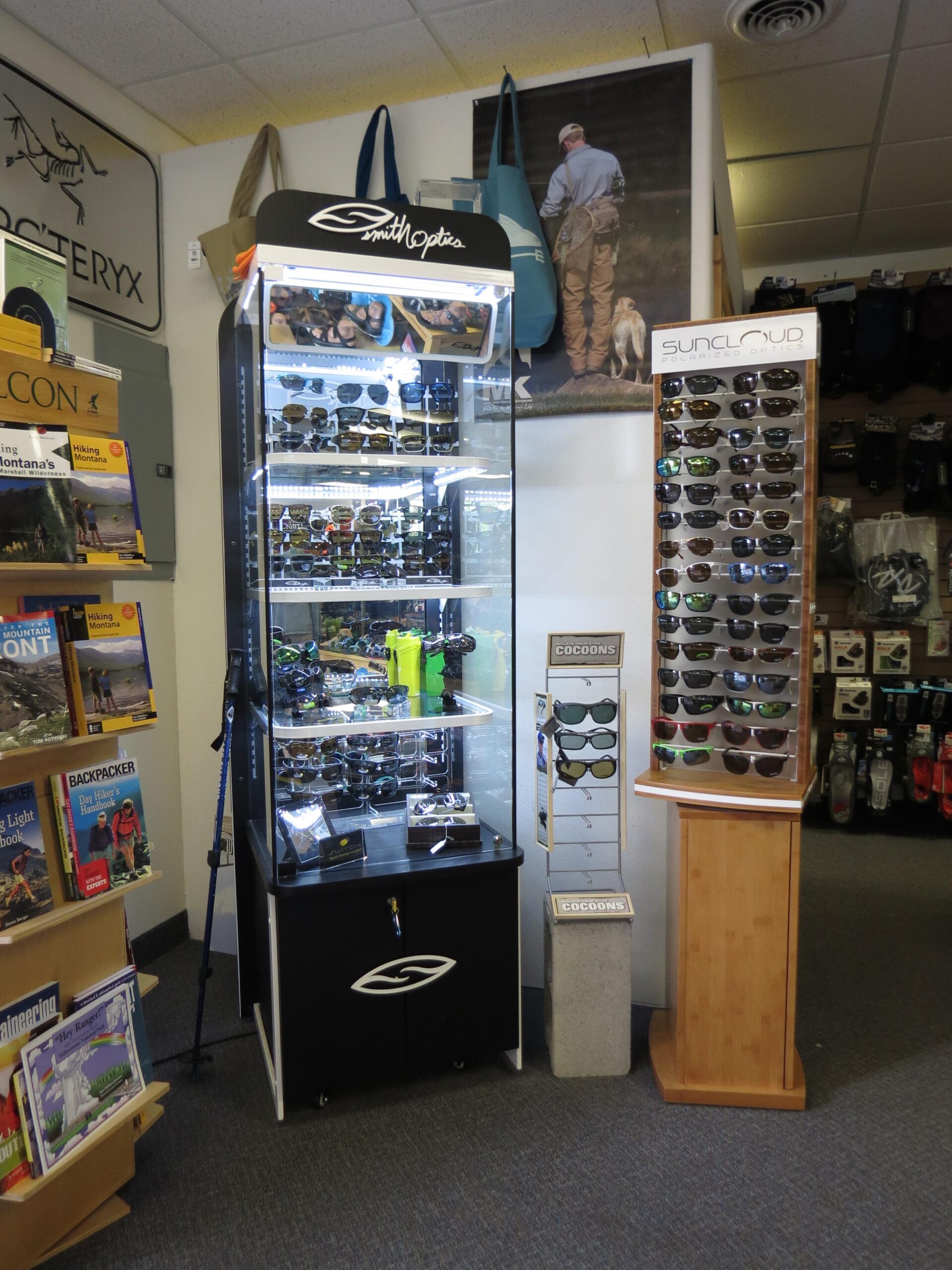 Display of sunglasses, featuring a glass showcase for Smith Optics on the left, and a wooden stand for SunCloud polarized optics on the right. Various books are visible on a shelf in the foreground, with outdoor gear in the background. The shop has a cozy, adventure-themed atmosphere.