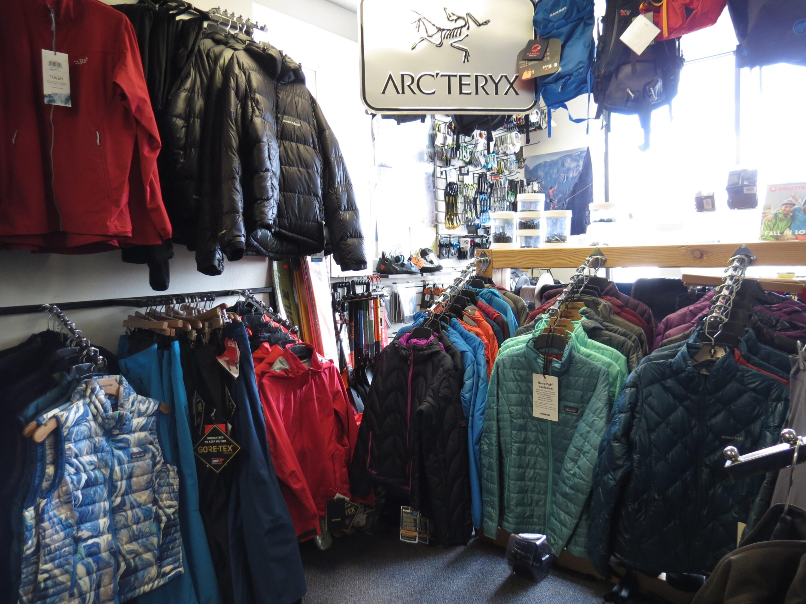A display of outdoor clothing in a retail store, featuring various styles of jackets and vests from brands like Arc'teryx and Patagonia. The garments include insulated jackets in bright colors, hanging racks filled with vests and outerwear, and a selection of gear accessories visible in the background. The store has a bright, organized atmosphere with sunlight coming through the windows.