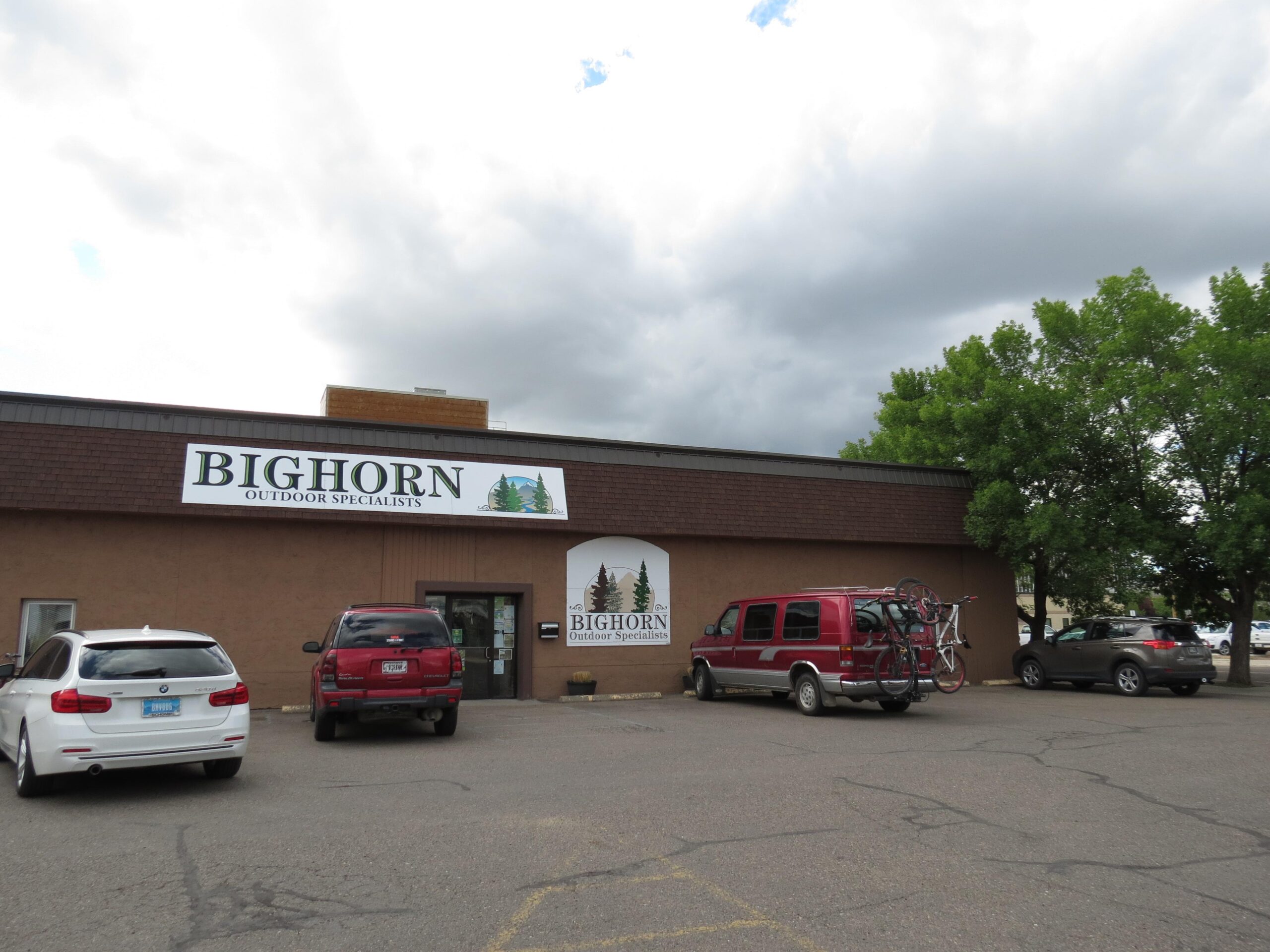Image of a store front with a large sign that reads "BIGHORN OUTDOOR SPECIALISTS." The building is brown with a shingled roof, and there are several vehicles parked in front, including a white car, a red van with bicycles on the back, and a dark-colored SUV. In the background, there are trees and a cloudy sky.