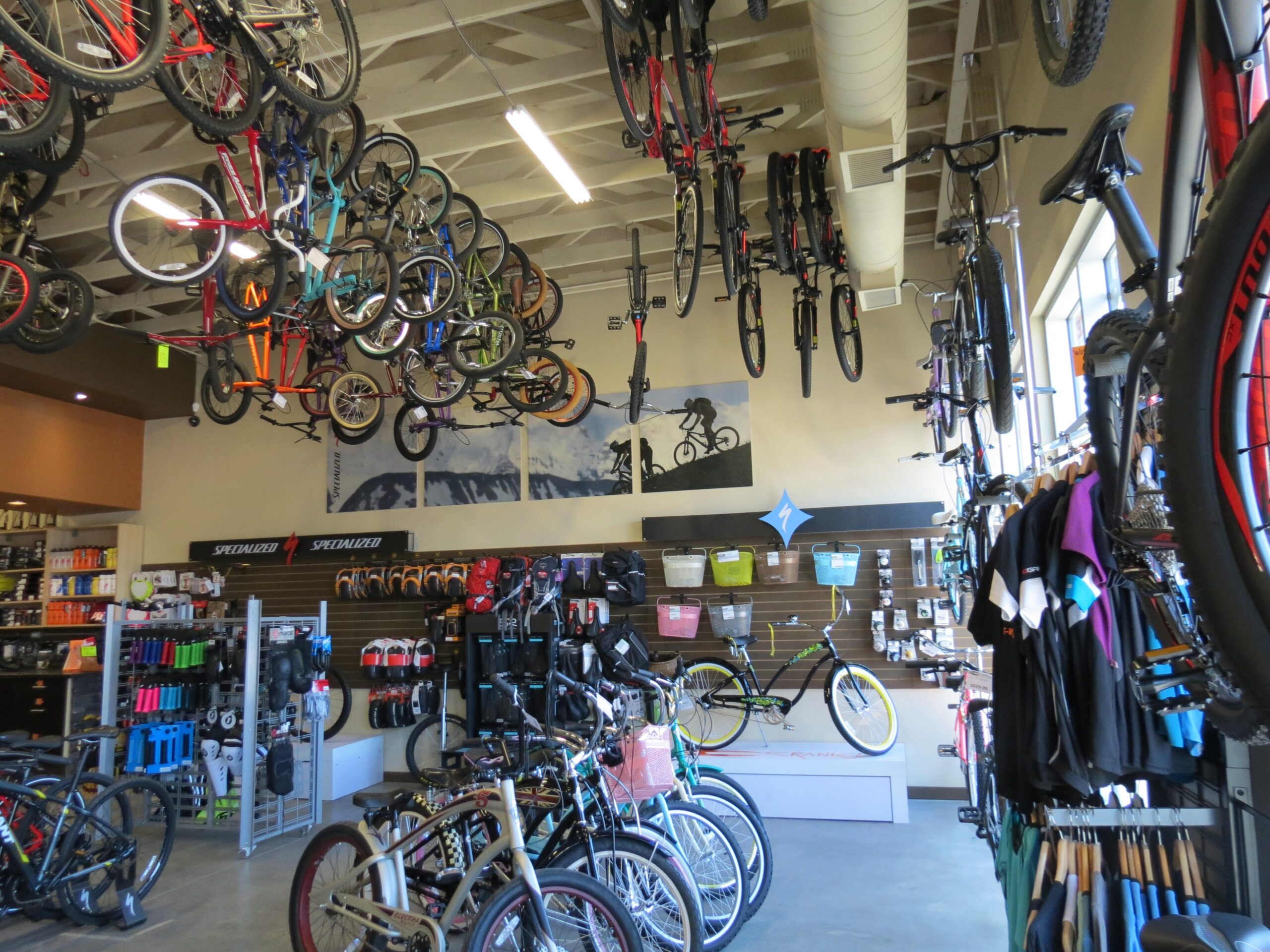A vibrant indoor bicycle shop displaying a variety of bikes hanging from the ceiling and arranged on the floor. The walls feature cycling apparel and accessories, with shelves filled with helmets and gear. Bright lighting and a mural of cyclists add energy to the space.