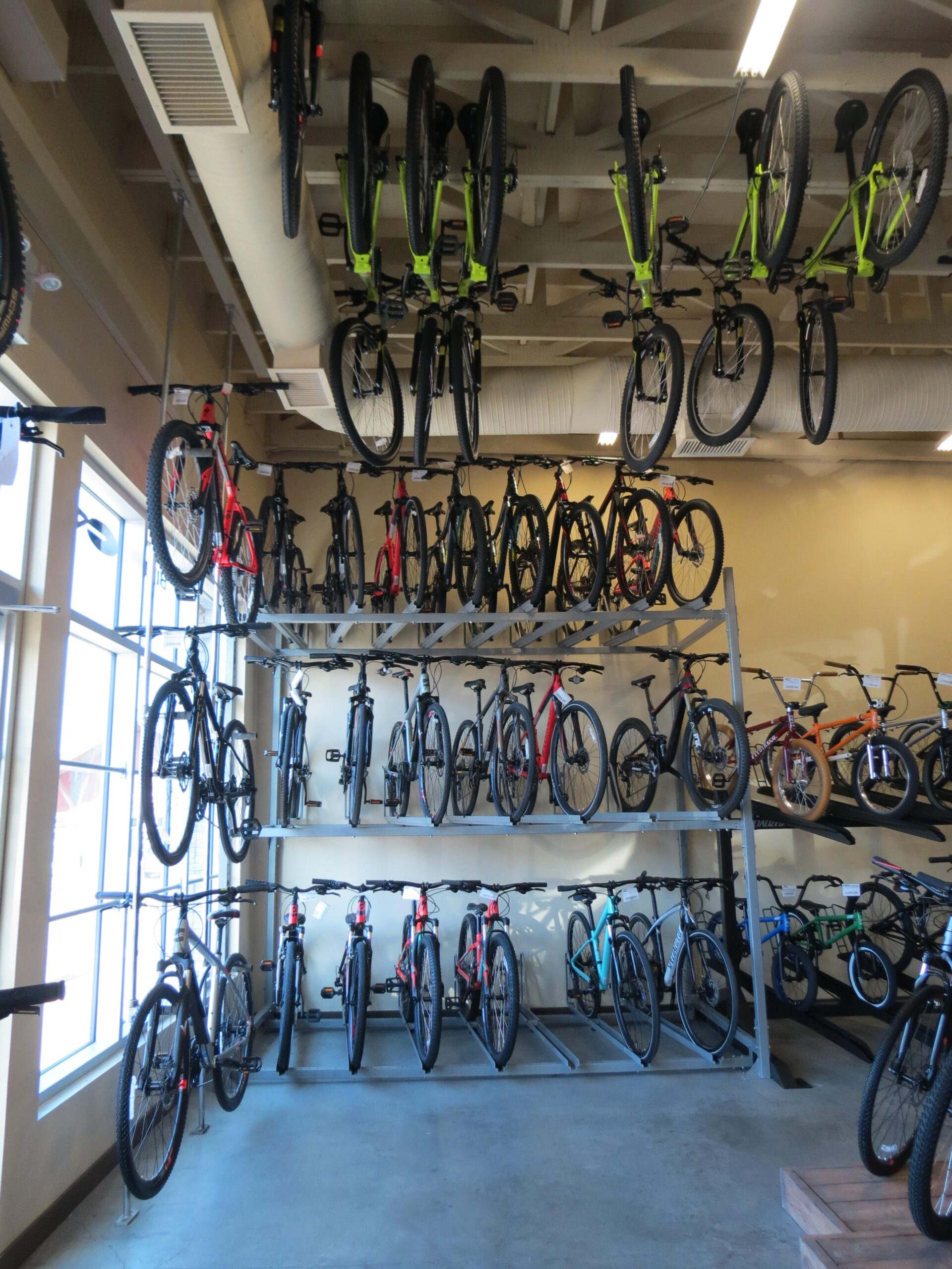 Image of a bicycle shop interior featuring a variety of bikes displayed both on the walls and on shelves. The bikes include several mountain bikes in various colors, including red, green, and black, with some bikes hanging upside down from the ceiling. The shop has a modern design with large windows letting in natural light, highlighting the bike selection.