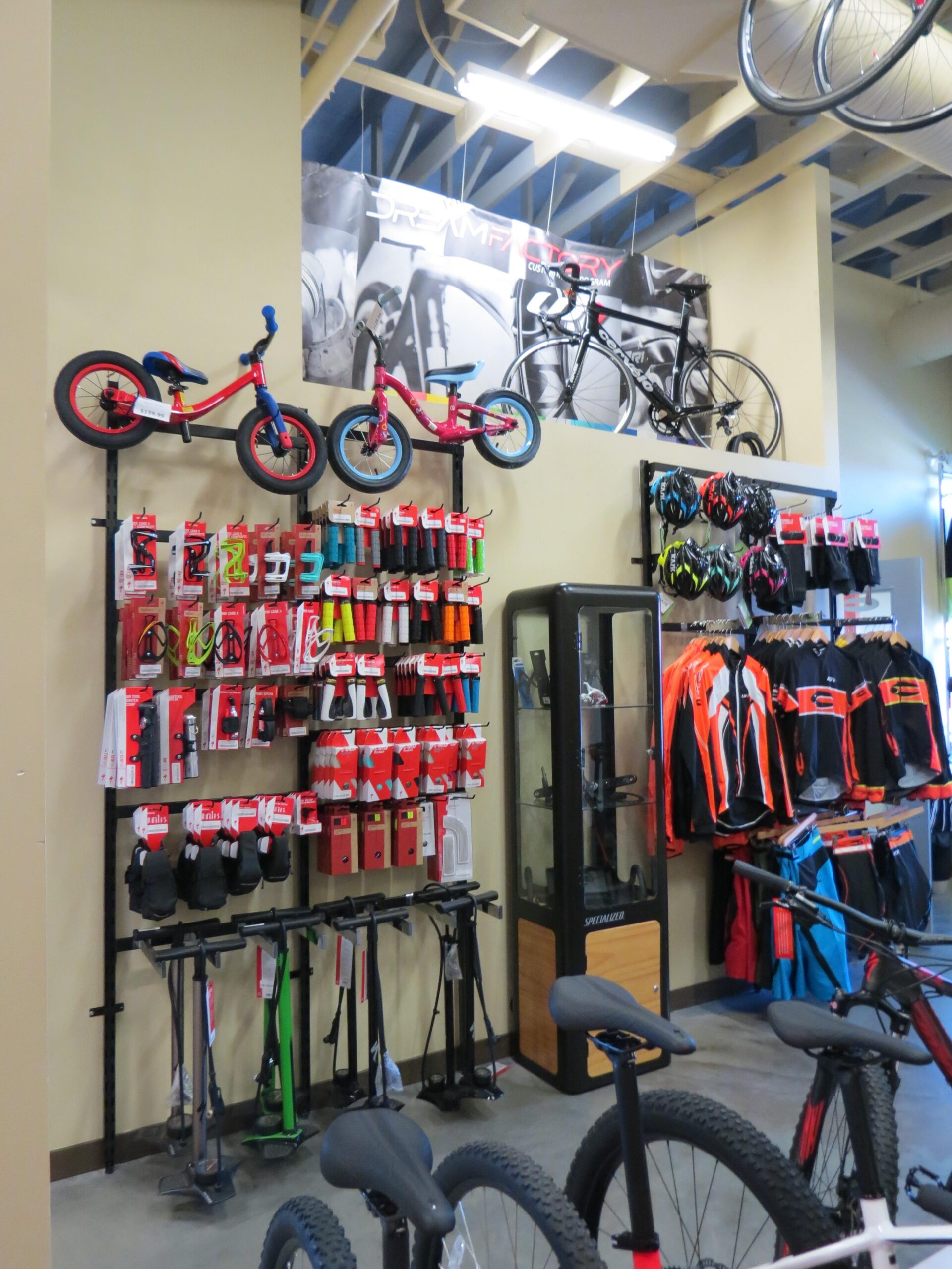 Image of a bicycle store interior showcasing various bicycles and biking accessories. The left side features colorful children's bicycles mounted on a wall display, while the lower section includes a rack filled with bike grips, tools, and other accessories. A glass display case is visible, containing more biking items. The right side showcases cycling apparel hanging on racks, with bright colors and designs, and biking helmets neatly arranged above. The store has a vibrant and organized layout, with a promotional banner in the background.