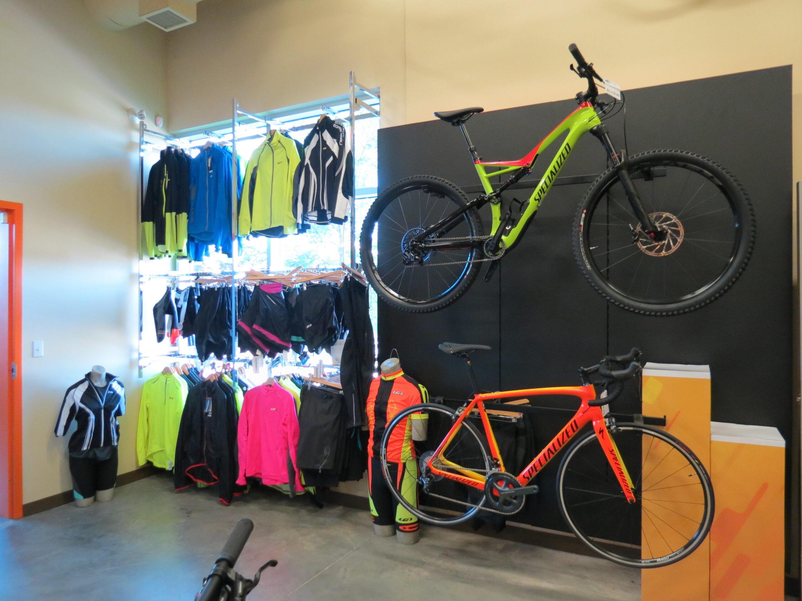 A display in a cycling shop featuring a variety of activewear including jackets and shorts on hanging racks. Two bicycles, one bright green and black mountain bike and one orange and black road bike, are mounted on the wall. The shop has a modern design with a concrete floor and orange accents.