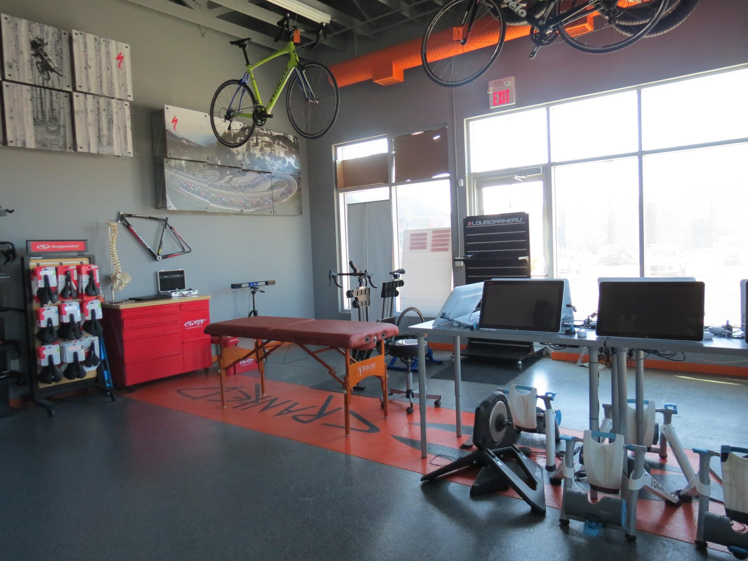 A bright indoor cycling studio with an active atmosphere. The space features a green bicycle hanging from the ceiling, large windows allowing natural light, and a variety of equipment including exercise machines, a therapy table, and monitoring screens. The walls display cycling-related artwork, and there are rows of cycling gear neatly organized on racks.