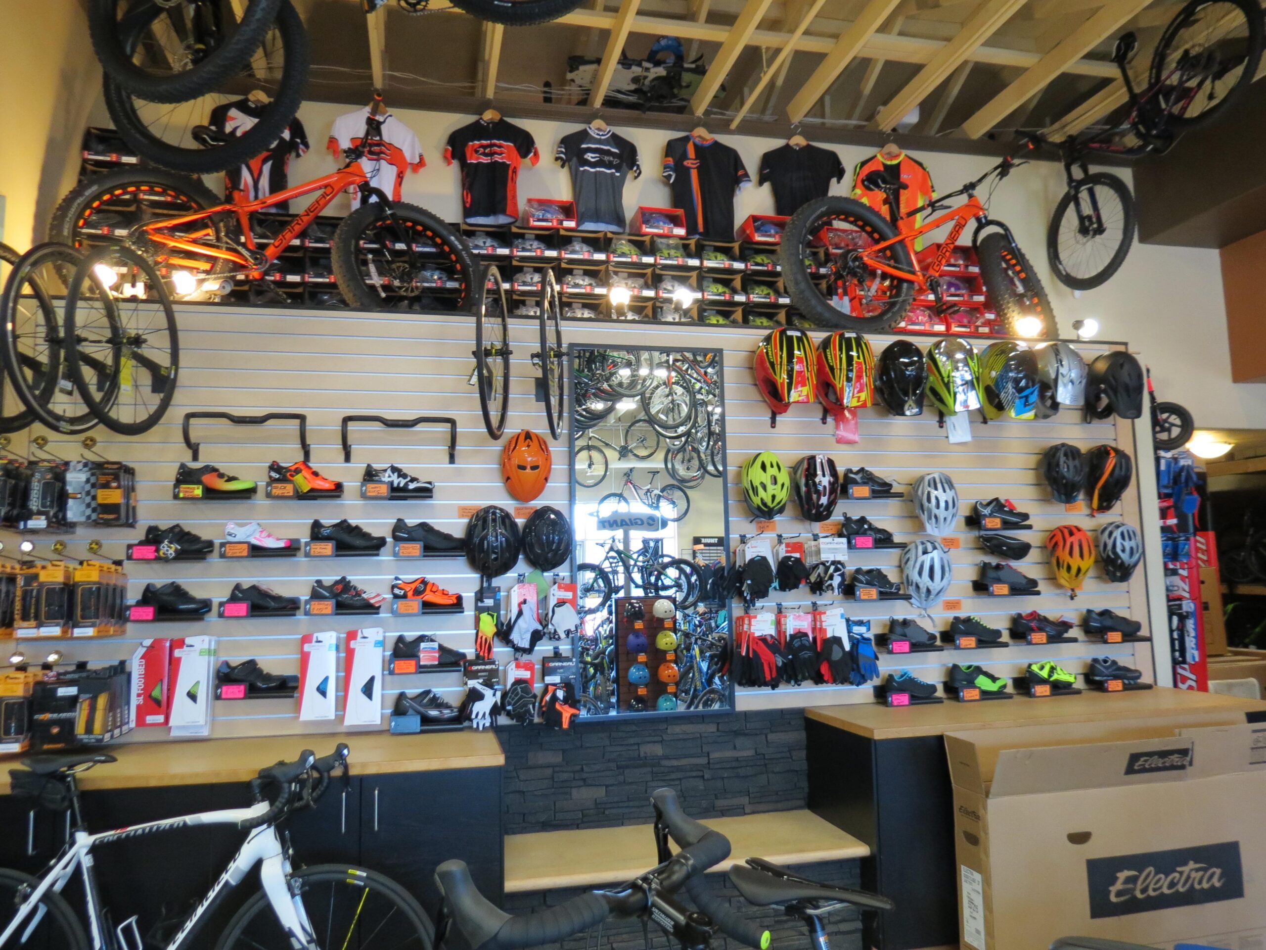 Interior of a bike shop displaying a variety of bicycles, helmets, and cycling shoes. The wall features several mountain bikes and road bikes hanging above a selection of helmet types. Below, cycling shoes and accessories are arranged neatly on shelves, with some items marked with price tags. The store has a modern and organized layout.