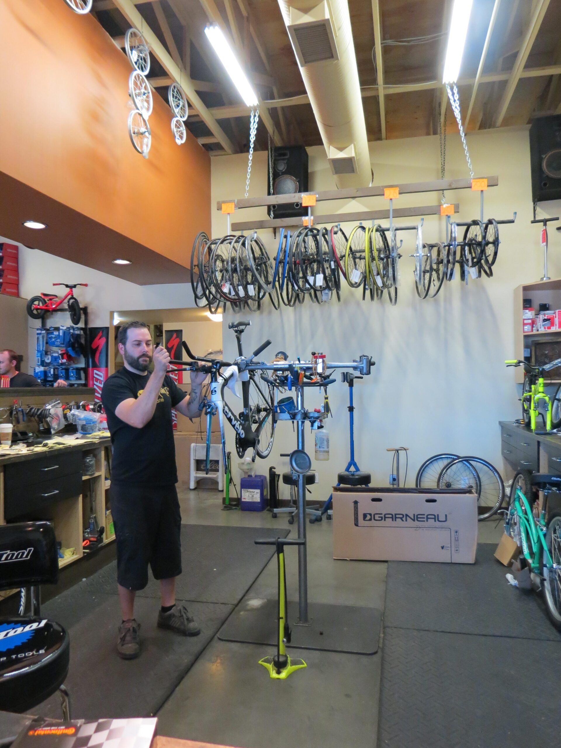 A bicycle mechanic is working in a bike shop, adjusting a bike on a repair stand. The shop features a variety of bike wheels hanging on the wall and tools arranged on a workbench. Boxes and completed bicycles are visible in the background.