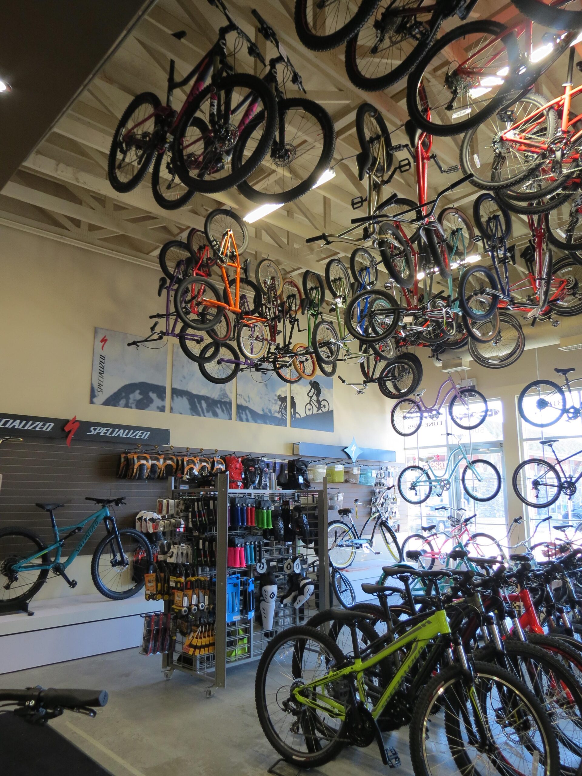 A brightly lit bicycle store featuring a variety of bicycles displayed on the ceiling and walls. The interior showcases several bike models in different colors, including mountain and road bikes, alongside a rack displaying biking accessories such as helmets, water bottles, and tools. The store has a modern feel with wooden beams and large windows, allowing natural light to brighten the space.