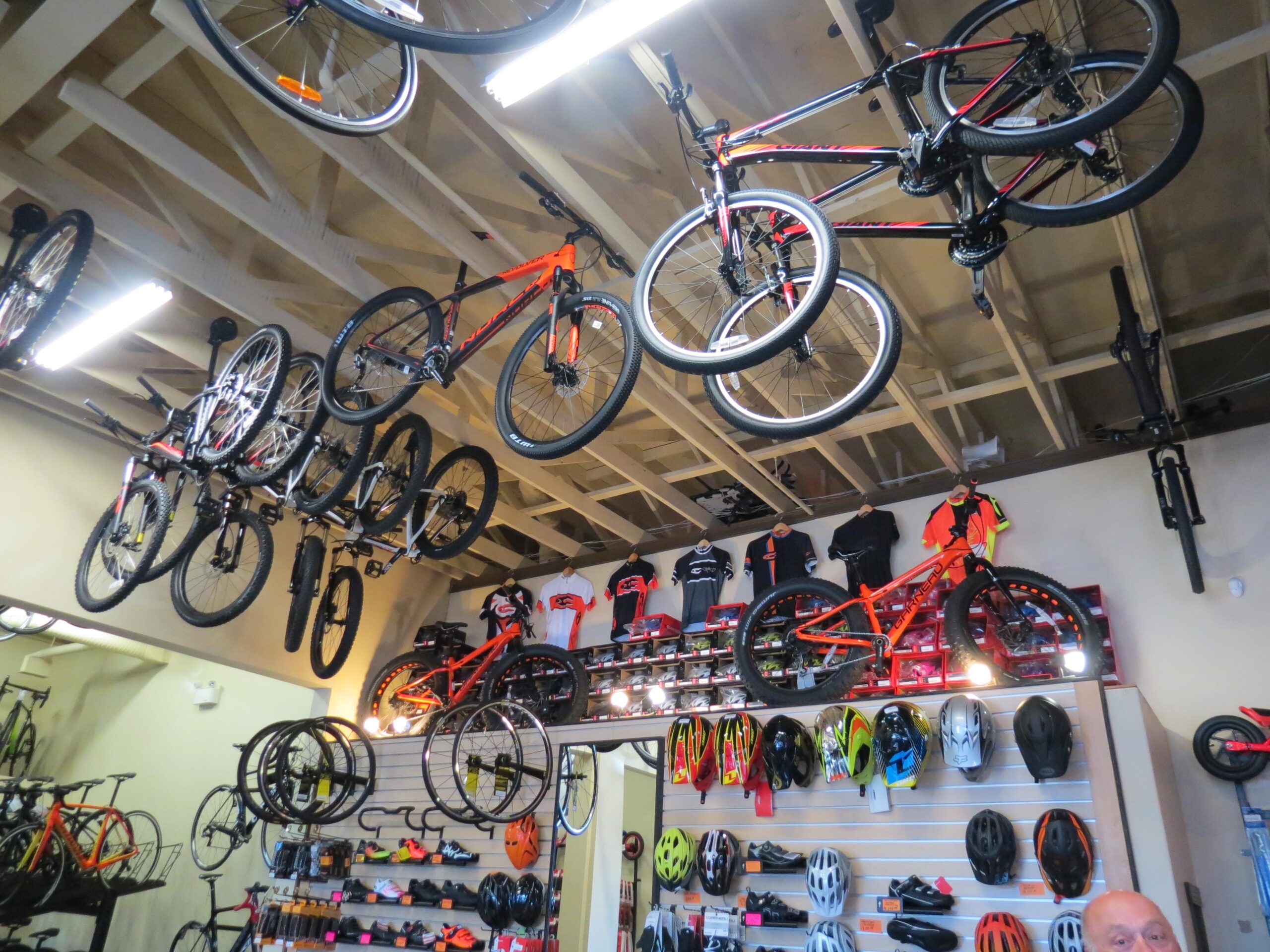 A bicycle shop interior featuring a variety of bicycles hanging from the ceiling and displayed on walls. The scene includes brightly colored mountain bikes, bicycle wheels, and an assortment of cycling apparel and helmets neatly arranged on shelves. The shop has a well-lit, organized layout, emphasizing a selection of biking gear.